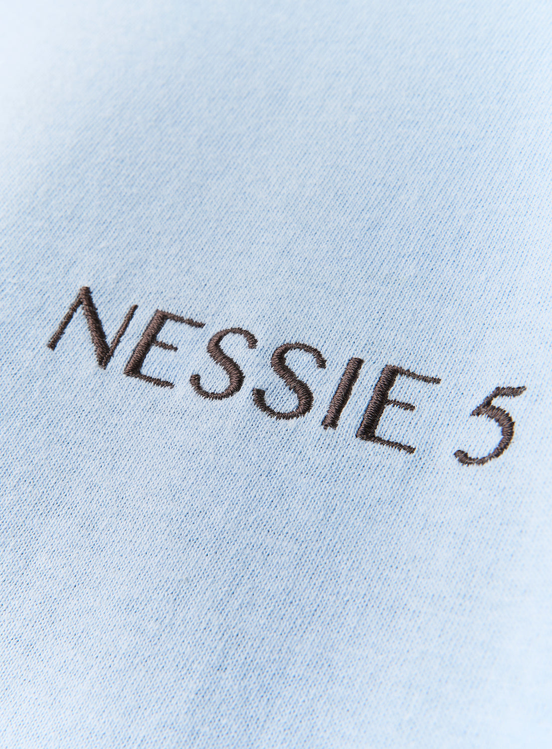Close-up of light blue fabric from the DAISAK Nessie T-Shirt, featuring the words NESSIE 5 embroidered in black thread.