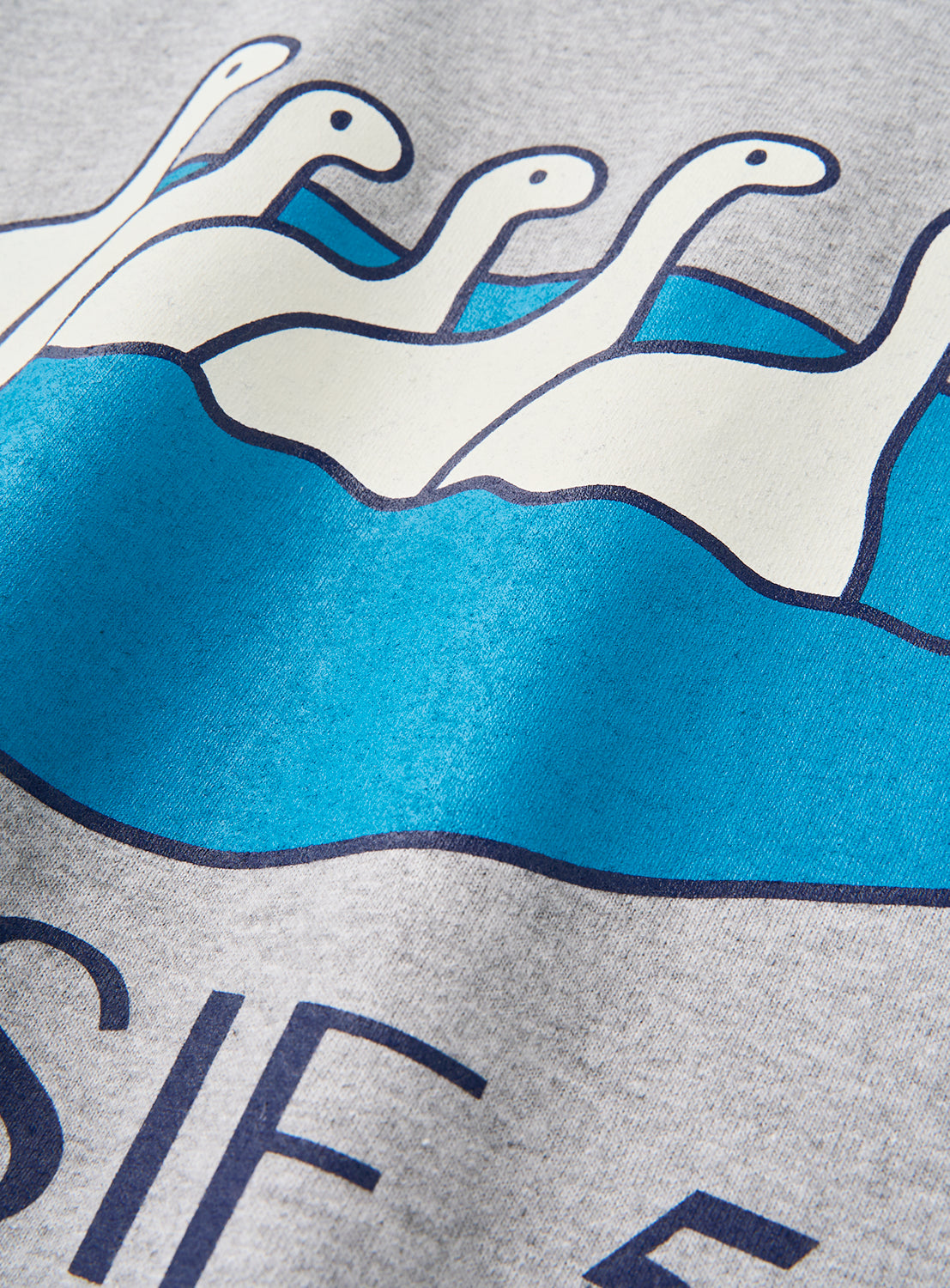 The DAISAK Nessie T-Shirt by DAISAK features a cartoon illustration of five white, long-necked Nessie-like creatures swimming in blue water, printed on a gray relaxed-fit graphic tee. Partial black text appears near the classic crew neckline at the bottom of the design.