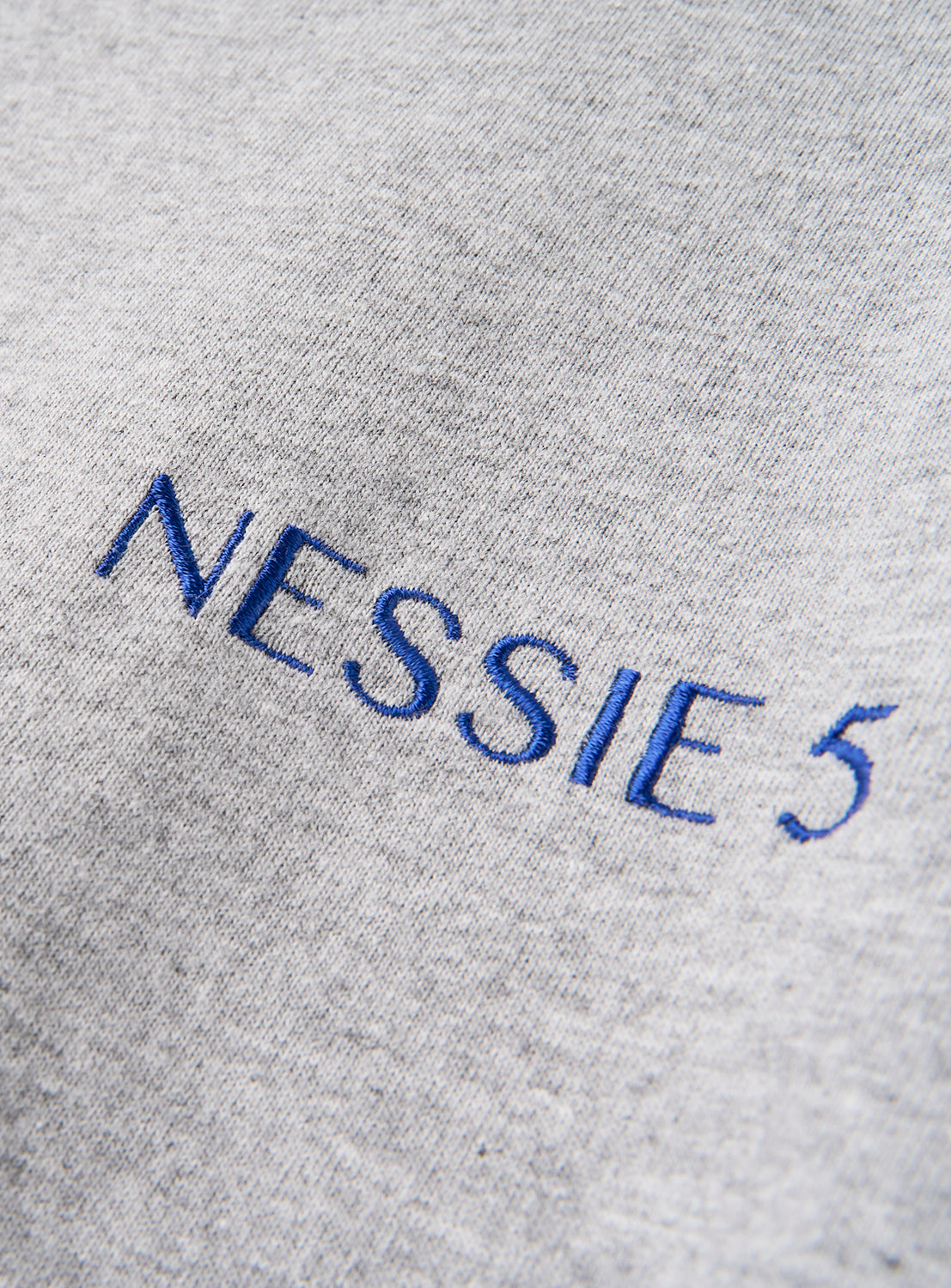 Close-up of the DAISAK Nessie T-Shirt shows gray fabric featuring blue, uppercase embroidery spelling out "NESSIE 5." The bold text pops against the textured material, giving this DAISAK tee a relaxed-fit look with classic graphic detailing.
