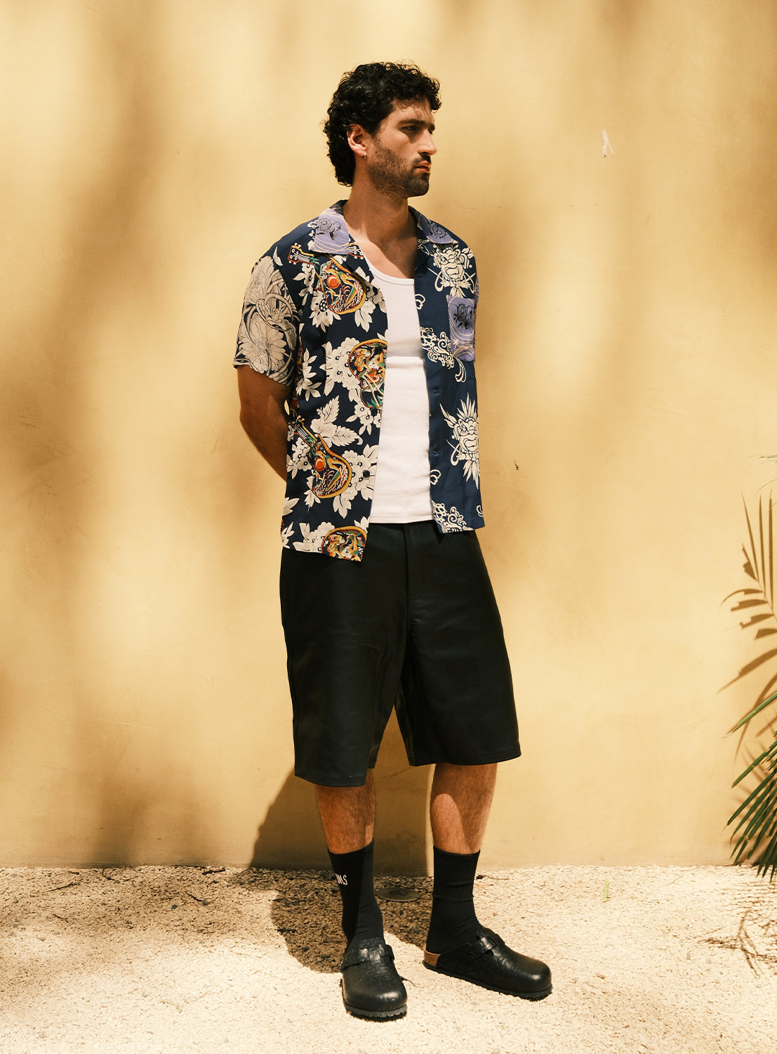 A man stands against a tan wall, wearing the SUN SURF Exclusive Patchwork Aloha Shirt 26SS by SUN SURF over a white tank top, paired with black shorts, black socks, and black slip-on shoes. His arms are behind his back as sunlight casts soft shadows.