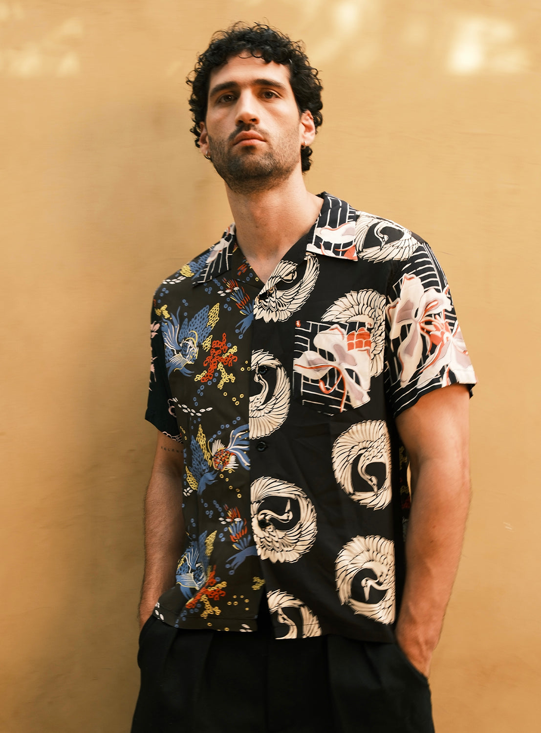 A man with curly dark hair stands against a plain tan background with his hands in his pockets, wearing the SUN SURF Exclusive Patchwork Aloha Shirt by SUN SURF—a short-sleeve shirt featuring bold, colorful patterns split down the middle.