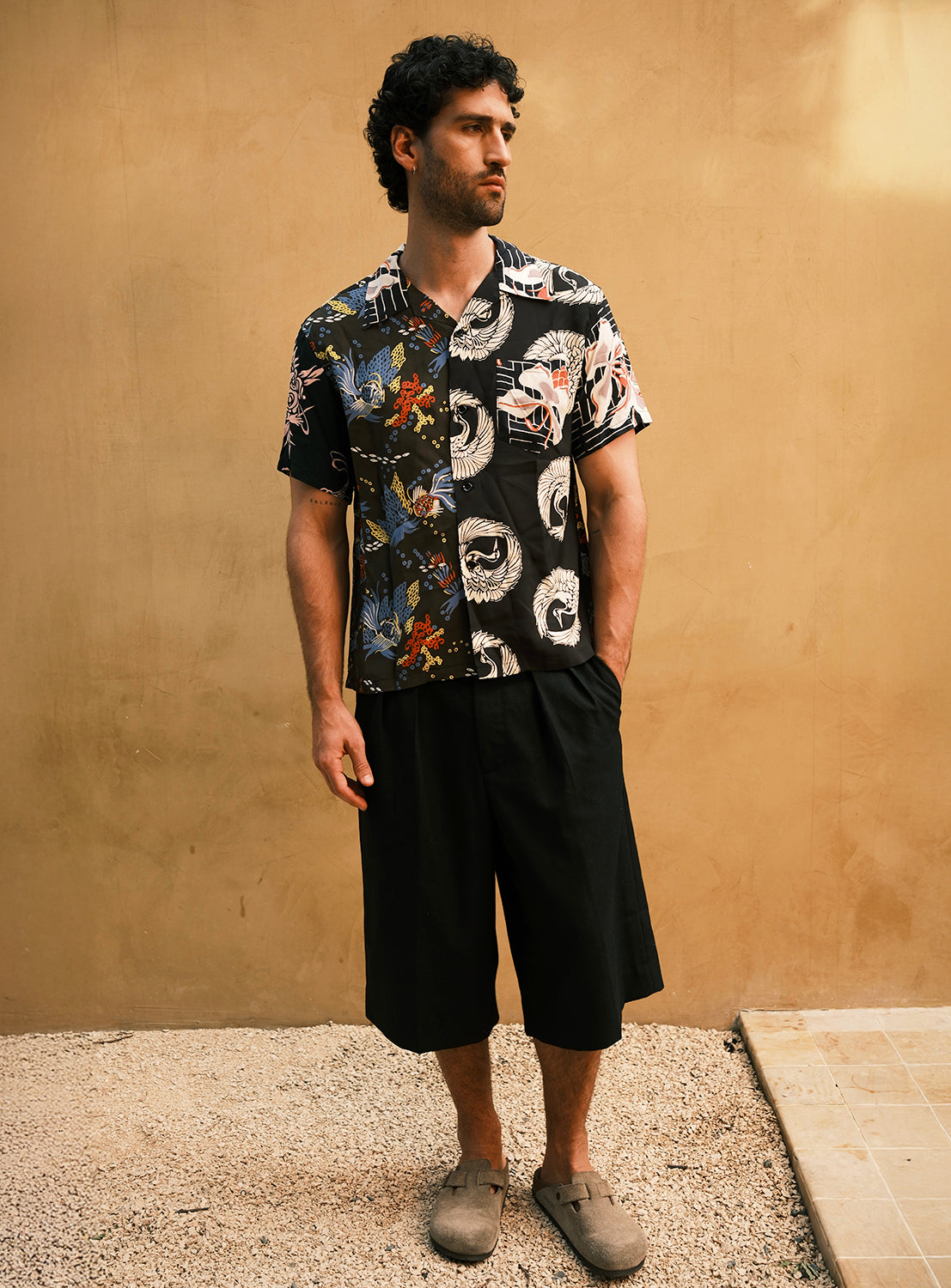 A man stands on gravel in front of a tan wall, wearing the SUN SURF Exclusive Patchwork Aloha Shirt by SUN SURF, paired with loose black shorts and brown slip-on shoes, looking to the side.