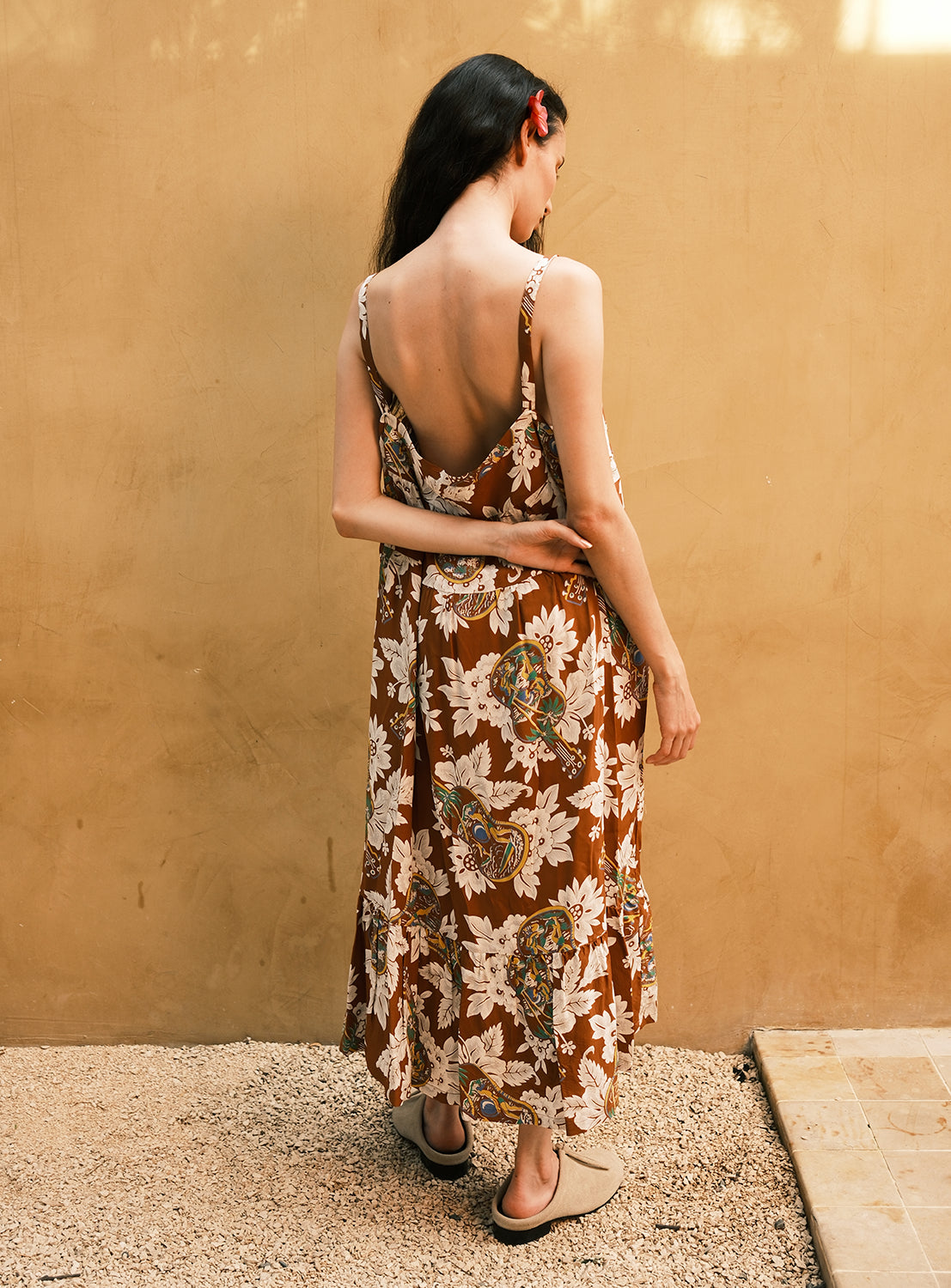 A woman with long dark hair and a red flower clip stands against a tan wall, facing away from the camera on a gravel surface, wearing the [Exclusive] SUN SURF Sleeveless Cami Dress by SUN SURF with an open back and black sandals.