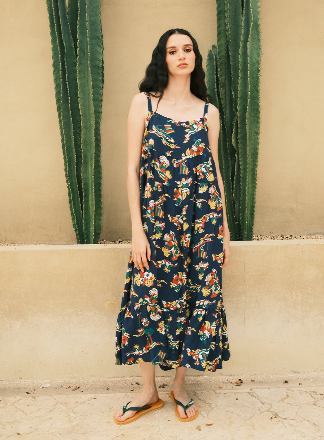 A woman wearing the [Exclusive] SUN SURF Sleeveless Cami Dress by SUN SURF—a dark floral sundress inspired by vintage aloha shirt dress styles—with sandals stands in front of a beige wall beside two tall green cacti.