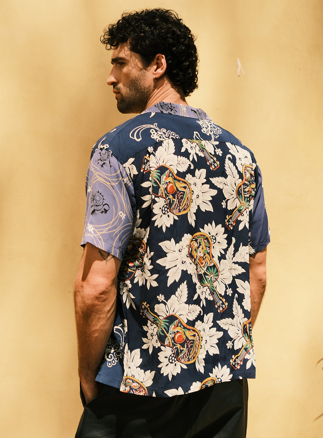 A man with curly dark hair, seen from behind, wears the SUN SURF Exclusive Patchwork Aloha Shirt 26SS—a blue short-sleeve shirt by SUN SURF featuring a bold floral and artistic motif inspired by vintage aloha shirts—standing against a plain light beige background.