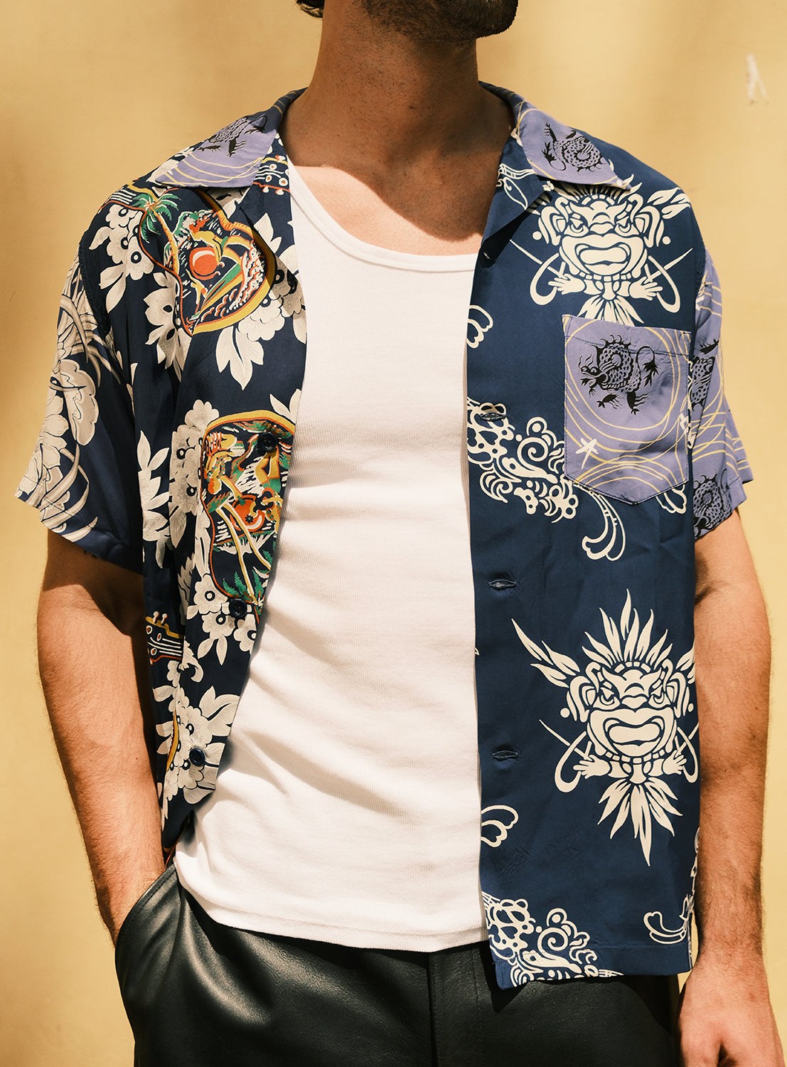 A person wearing the SUN SURF Exclusive Patchwork Aloha Shirt 26SS—a navy blue short-sleeve shirt by SUN SURF with white and colorful tiger and floral patterns inspired by vintage aloha shirts—layered over a white tank top and black pants, stands against a beige background.
