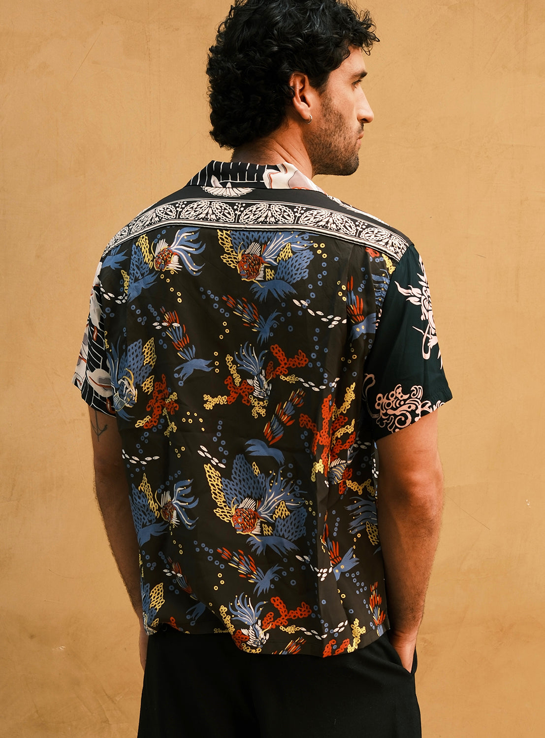 A man with short curly hair faces away, wearing the SUN SURF Exclusive Patchwork Aloha Shirt by SUN SURF, featuring intricate blue, red, yellow, and white designs reminiscent of vintage aloha shirts. He stands against a dark background in front of a plain beige wall.