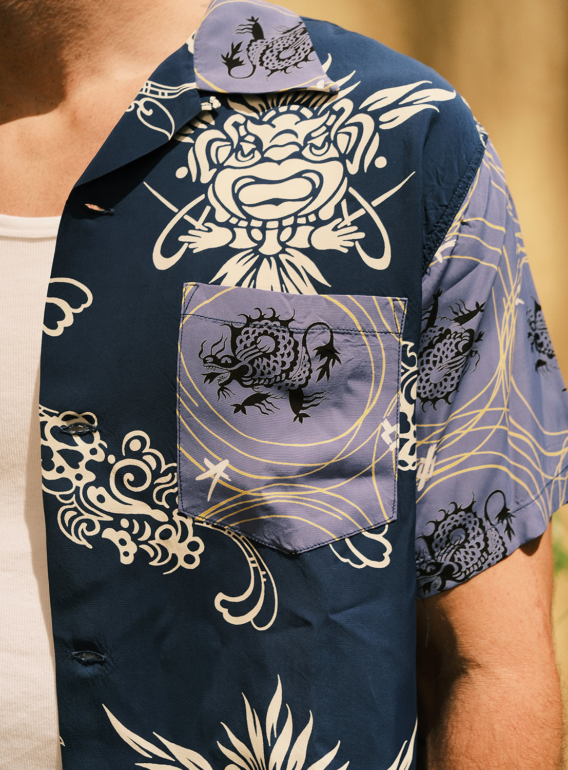 Close-up of a person wearing the SUN SURF Exclusive Patchwork Aloha Shirt 26SS—an eye-catching blue short-sleeve shirt by SUN SURF, featuring bold white and black dragon motifs with gold swirling accents over a white tank top. Inspired by vintage aloha shirts and crafted with original printing techniques, only the upper torso and part of the arm are visible.