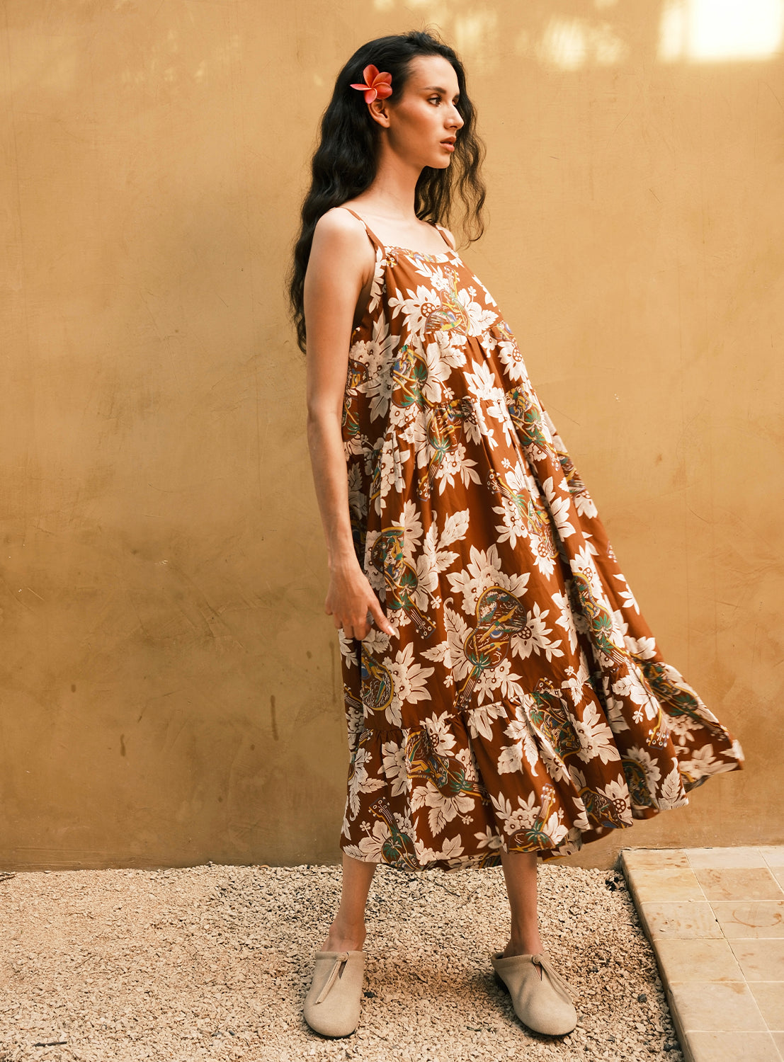 A woman stands sideways against a beige wall and pebbled ground, wearing the [Exclusive] SUN SURF Sleeveless Cami Dress by SUN SURF, styled with tan shoes and a red flower in her long dark hair.
