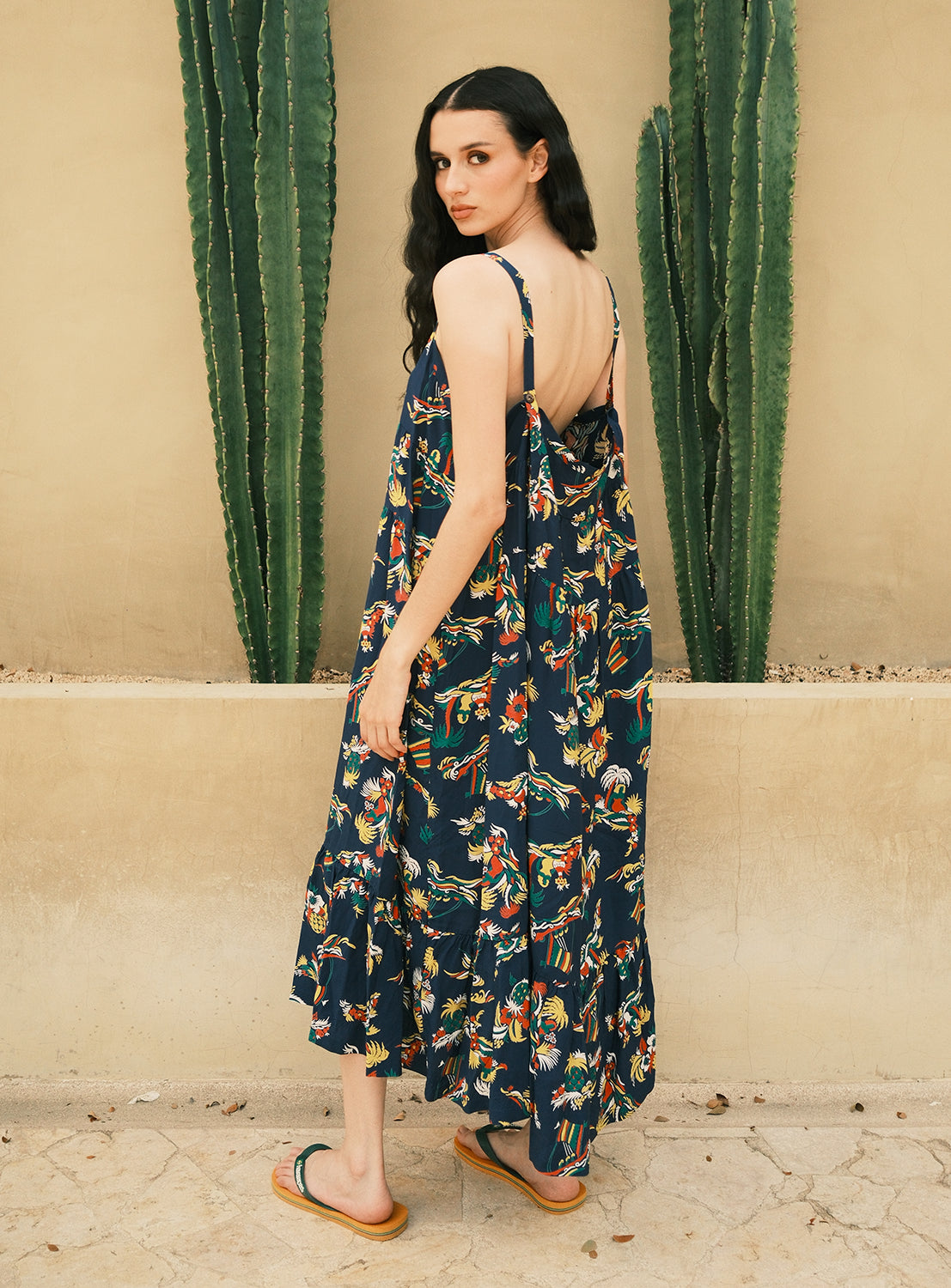 A woman wearing the [Exclusive] SUN SURF Sleeveless Cami Dress and sandals stands outdoors, her back turned but glancing over her shoulder. Two tall cacti rise behind her against a beige wall.