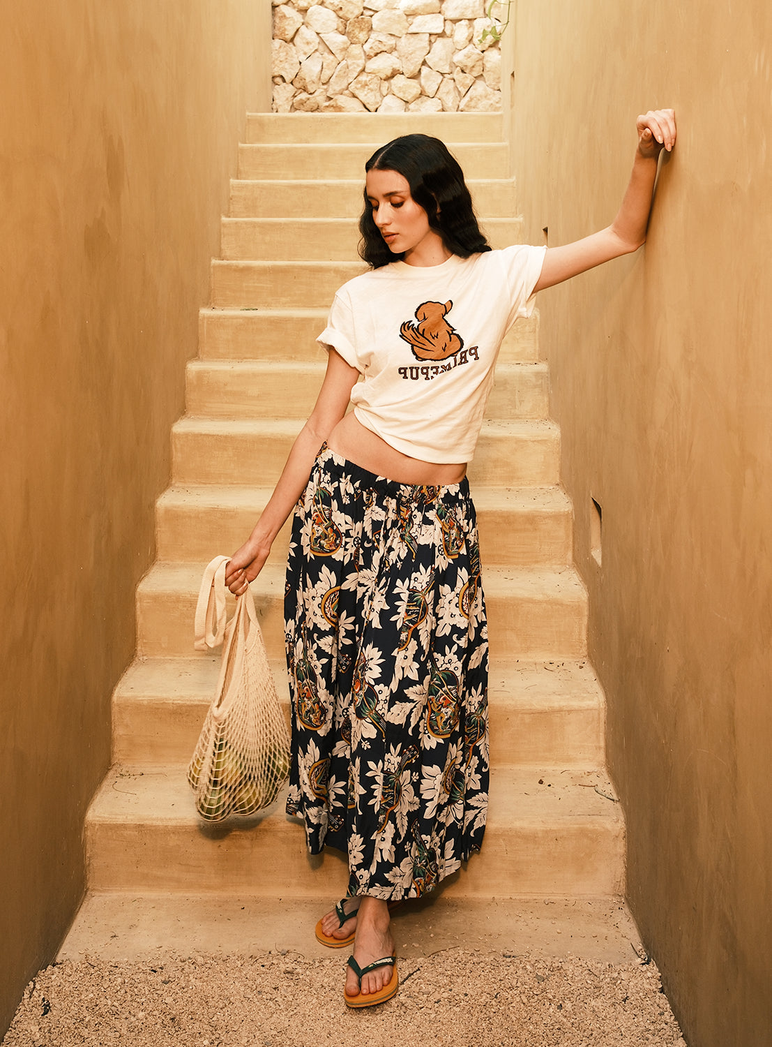 A woman stands on tan-colored stone steps, leaning against the wall. She wears a printed T-shirt, flip-flops, and holds a mesh bag with groceries. Her outfit features the [Exclusive] SUN SURF x BEAMS BOY Aloha Gathered Long Skirt by SUN SURF, showcasing vintage-inspired Hawaiian prints.