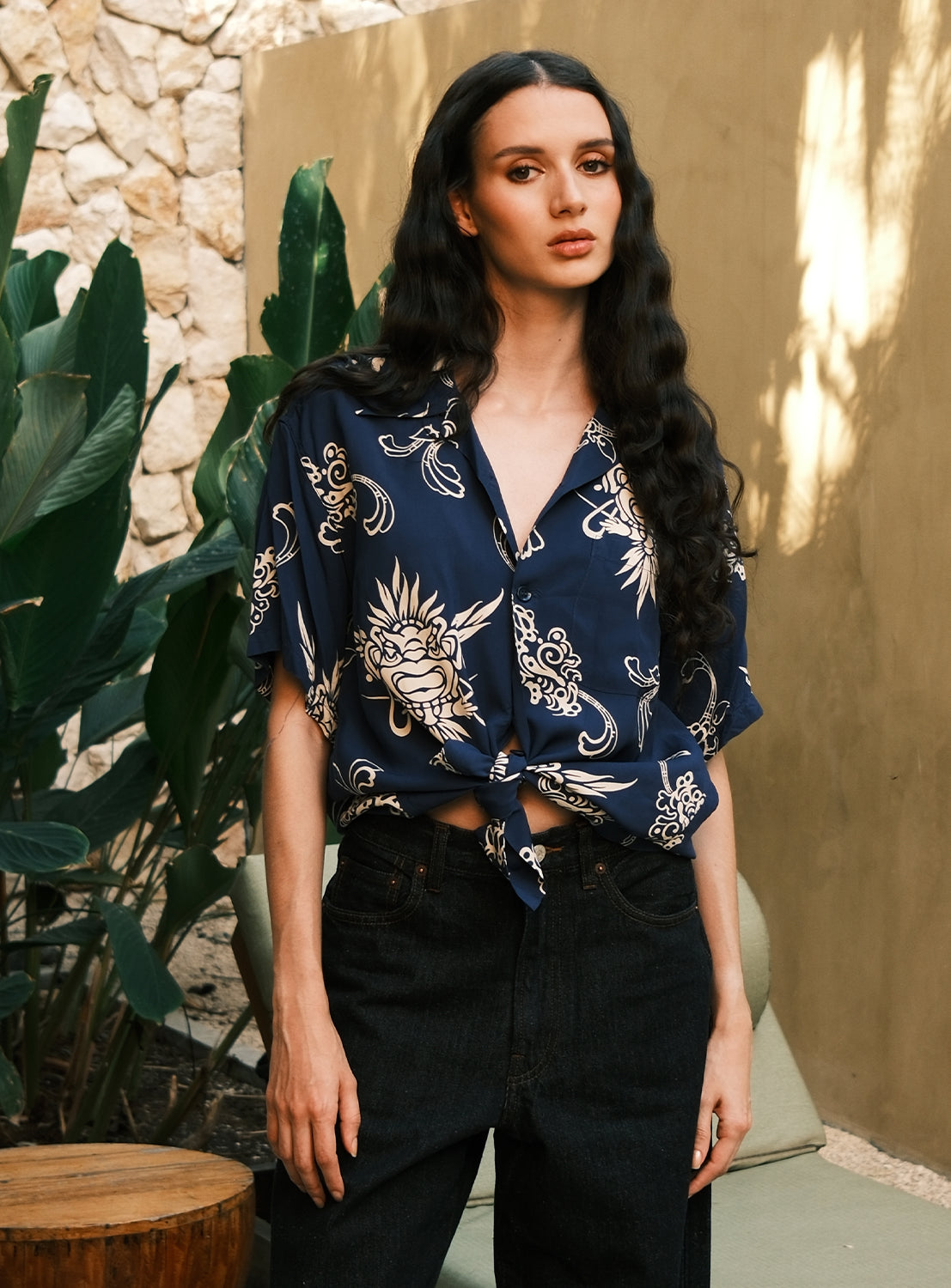A person with long dark hair stands by green plants, wearing the [Exclusive] SUN SURF x BEAMS BOY Open Collar Shirt from SUN SURF, styled tied at the waist with black jeans, in front of a stone wall and a beige background.
