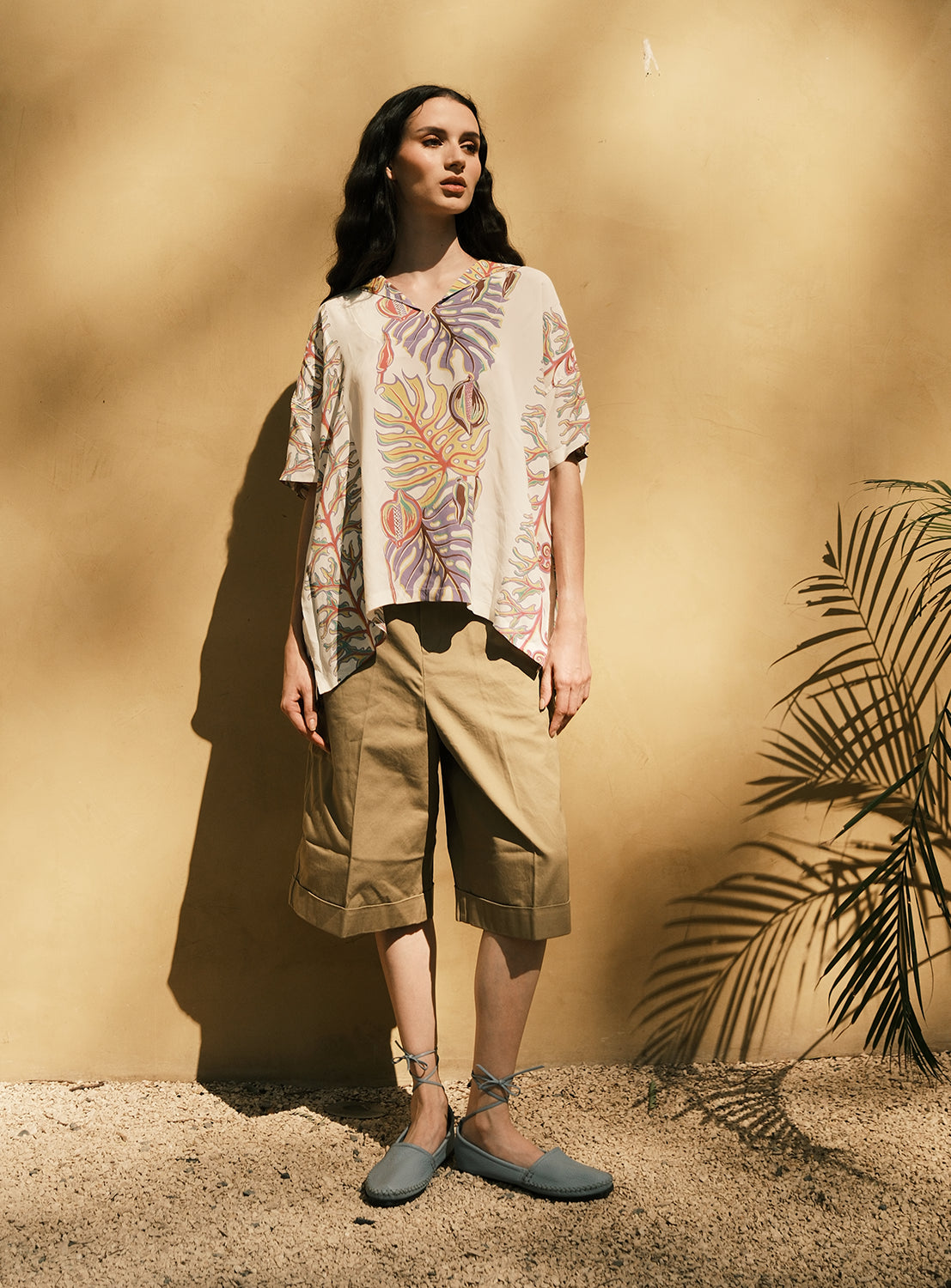 A person stands against a sunlit beige wall, wearing the [Exclusive] SUN SURF x BEAMS BOY Sailor Shirt by SUN SURF—a loose floral top inspired by vintage Hawaiian prints—paired with tan knee-length shorts and blue espadrilles. A plant casts a shadow on the ground and wall beside them, enhancing the laid-back SUN SURF × BEAMS BOY atmosphere.