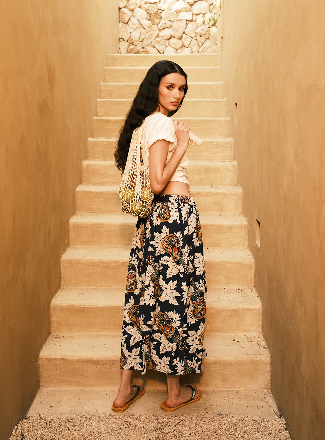 A woman with long dark hair stands on outdoor stone steps, wearing a light top, the [Exclusive] SUN SURF x BEAMS BOY Aloha Gathered Long Skirt from SUN SURF, flip-flops, and a netted bag over her shoulder as she looks back toward the camera.