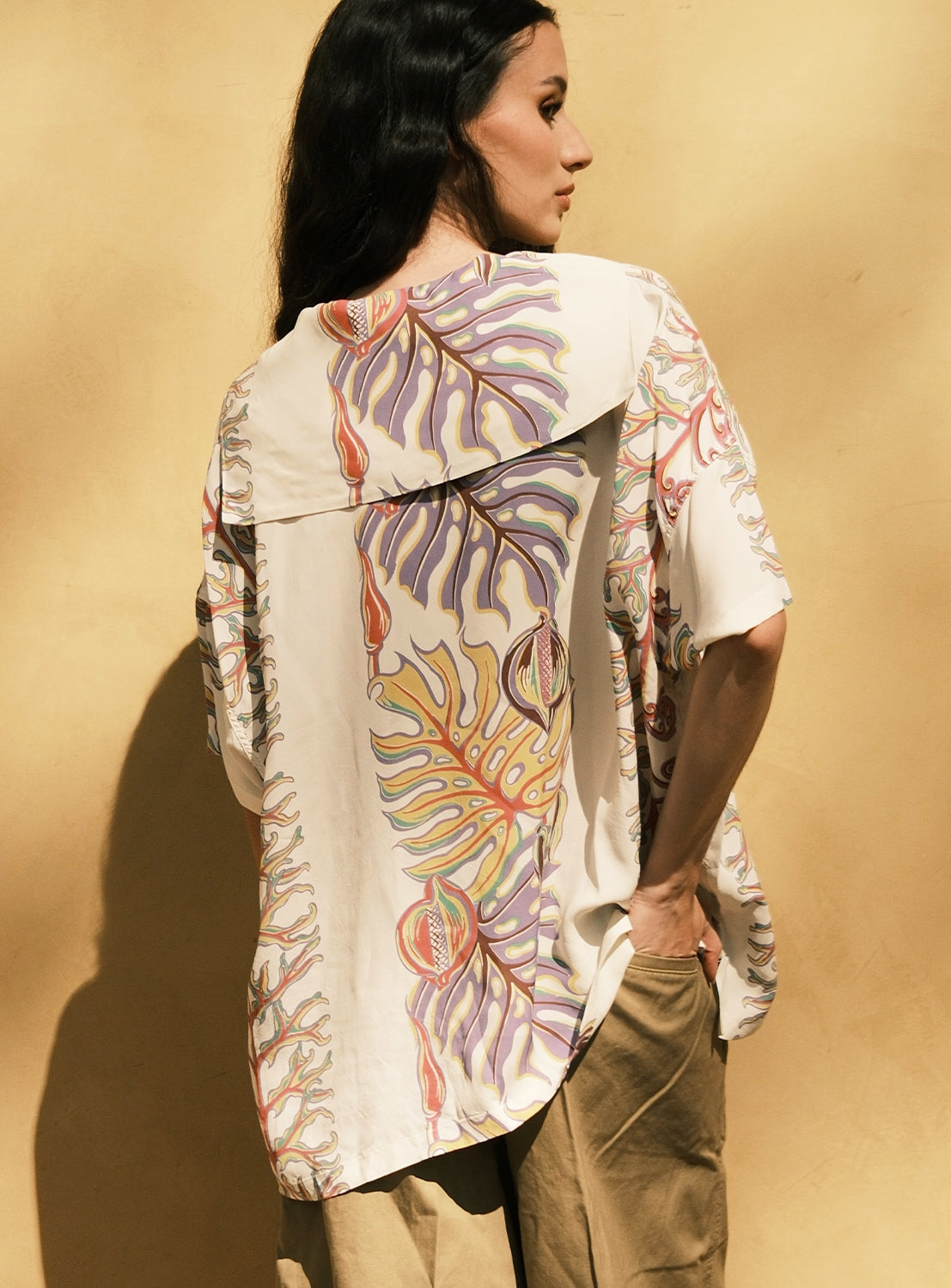 A woman stands against a yellow wall, facing away. She wears the [Exclusive] SUN SURF x BEAMS BOY Sailor Shirt from SUN SURF—a loose, cream-colored shirt featuring colorful botanical patterns inspired by vintage Hawaiian prints—and light brown pants, with one hand in her pocket.