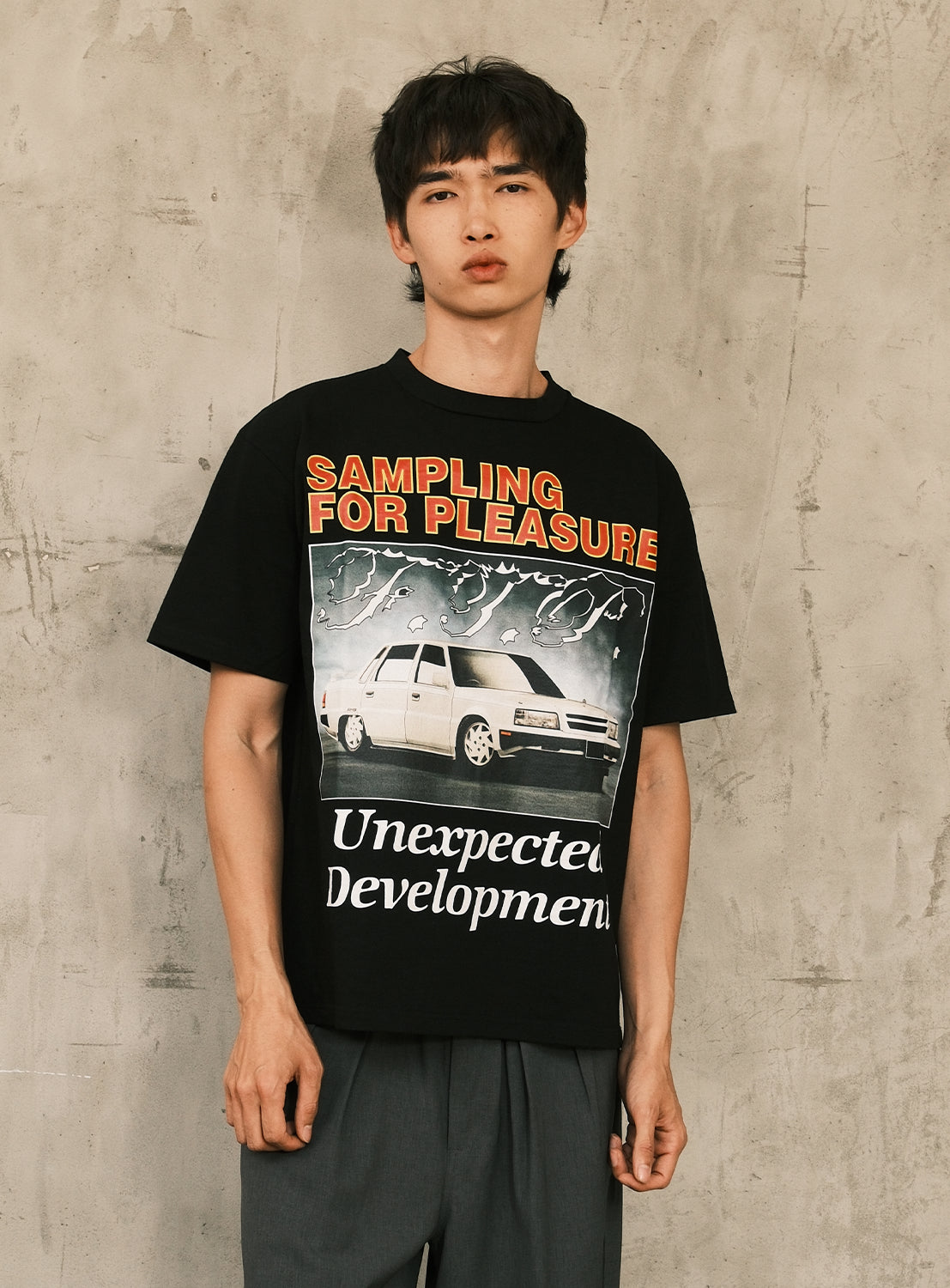 A person stands against a textured wall, wearing the BIAS DOGS Sampling For Pleasure Tee—a black graphic T-shirt featuring a striking car design and the text "SAMPLING FOR PLEASURE" and "Unexpected Development"—effortlessly channeling the bold style of vintage automotive shirts.