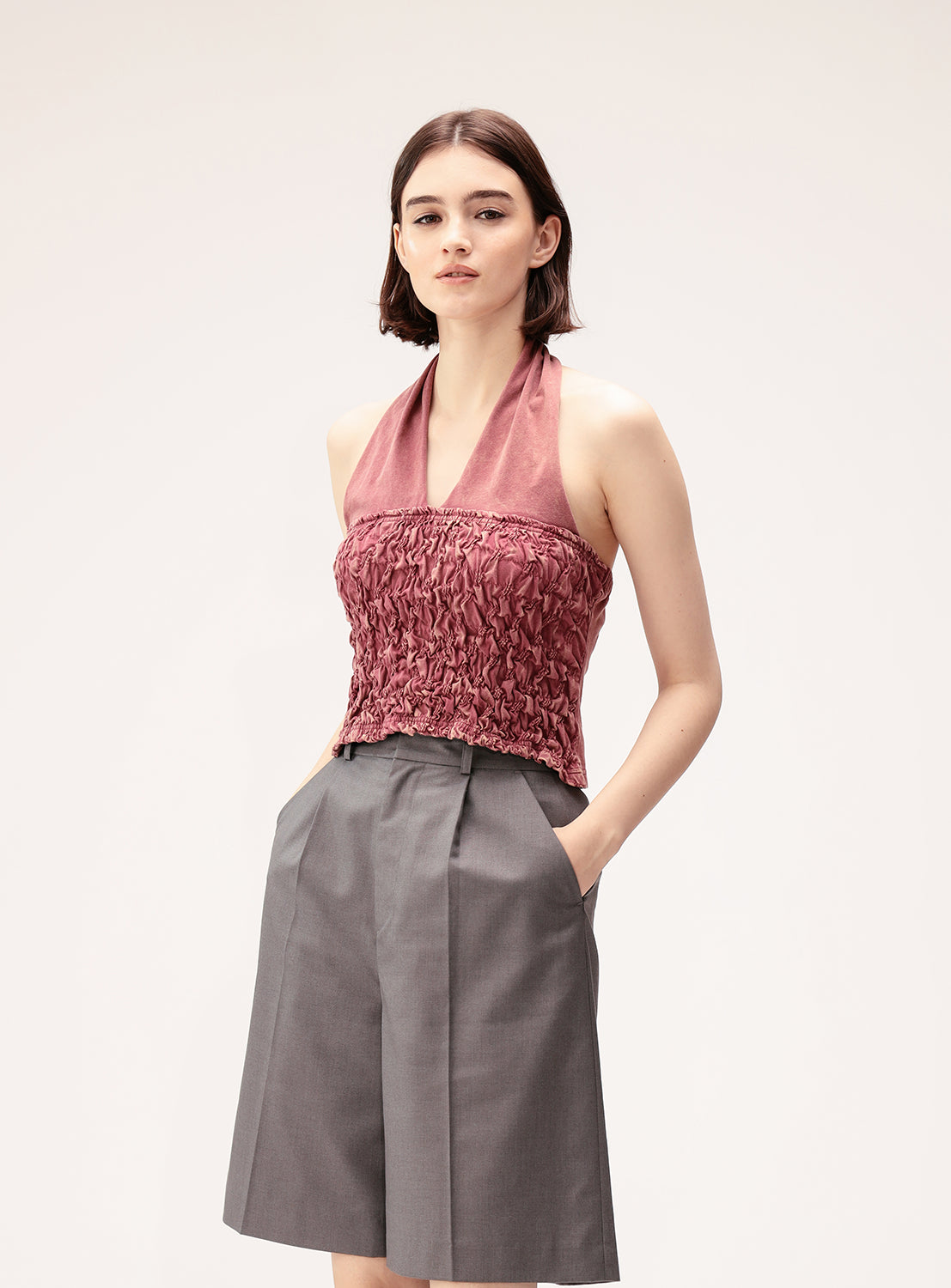 A person stands with hands in pockets, wearing the Ray BEAMS Shirred Halter Neck Top—a vintage-inspired, sleeveless mauve piece—paired with high-waisted gray knee-length shorts against a plain light background.