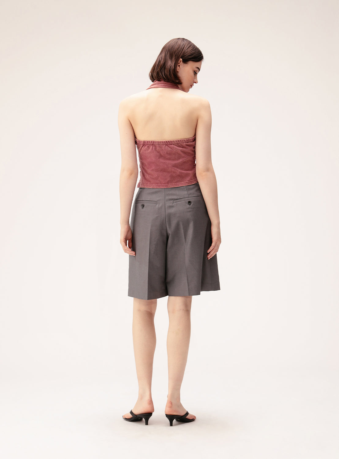 A woman stands facing away, wearing the Ray BEAMS Shirred Halter Neck Top in a vintage-inspired pink style, paired with gray knee-length shorts featuring back buttons and black heeled sandals, set against a plain light background.