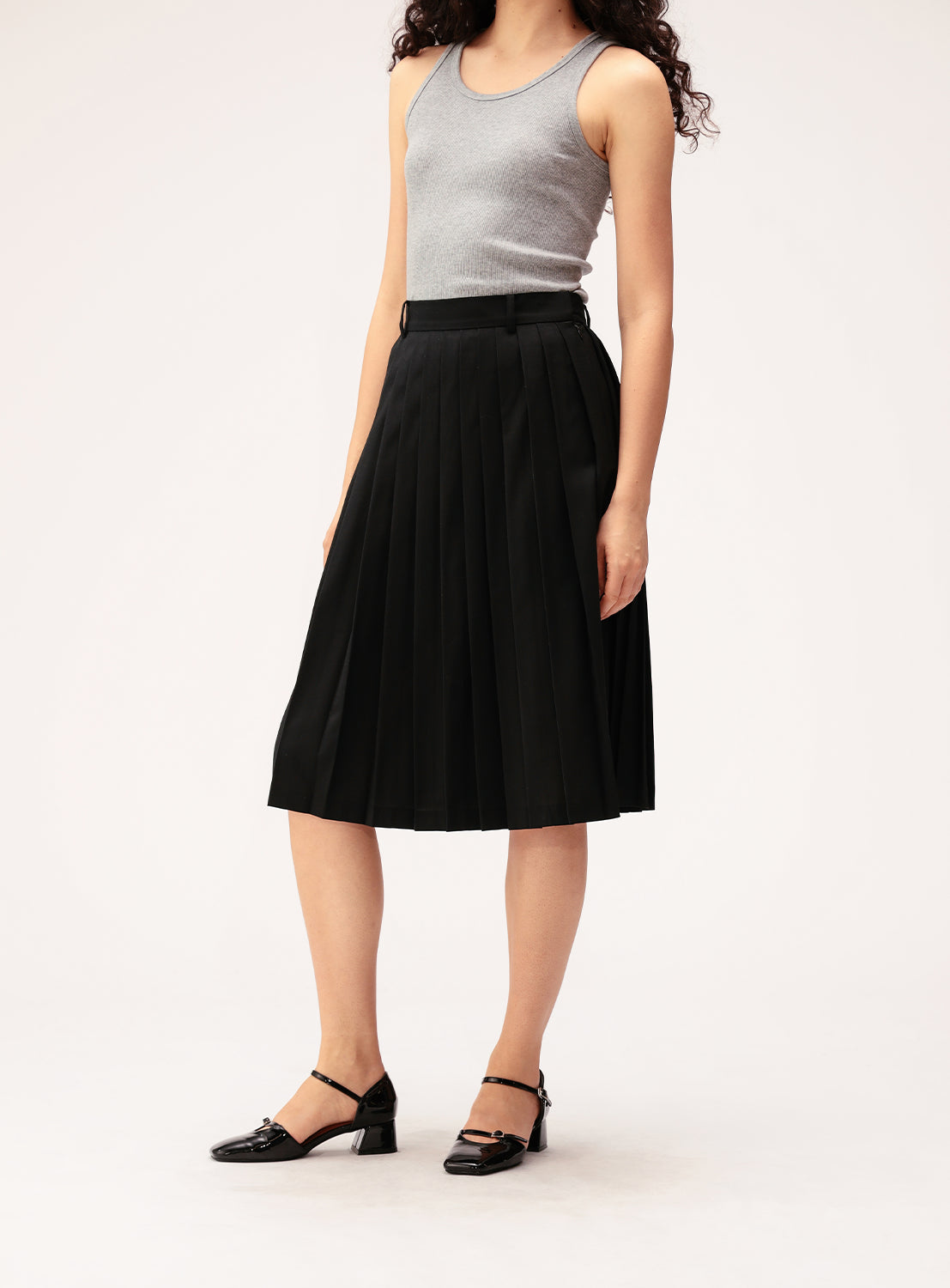 A woman stands on a plain light background wearing a gray sleeveless top, the BEAMS BOY Mixed Pleat Skirt—a black knee-length pleated skirt crafted from lightweight fabric—and black heeled Mary Jane shoes.