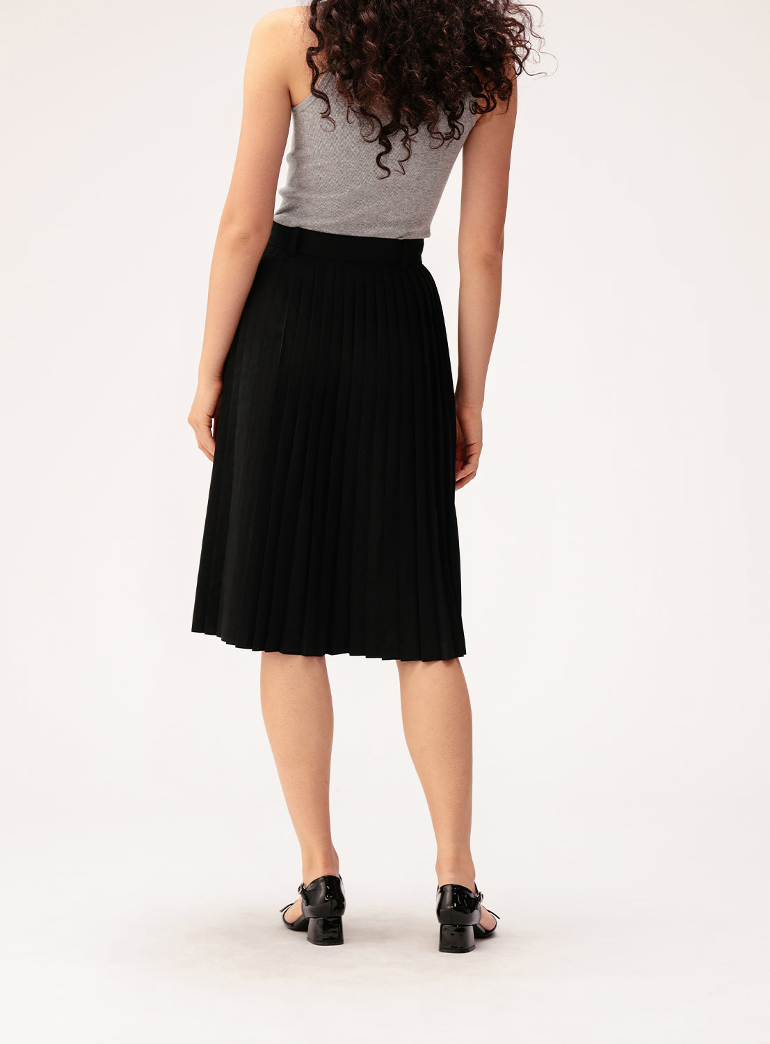 A person with curly hair, dressed in a gray sleeveless top, the BEAMS BOY Mixed Pleat Skirt—a black, knee-length skirt made from lightweight fabric—and black sandals stands facing away against a light background.