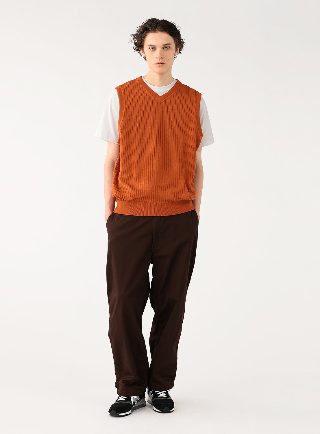 Model wearing a rust-colored ribbed knit vest over a white t-shirt and black pants, full front view, displaying casual layering.