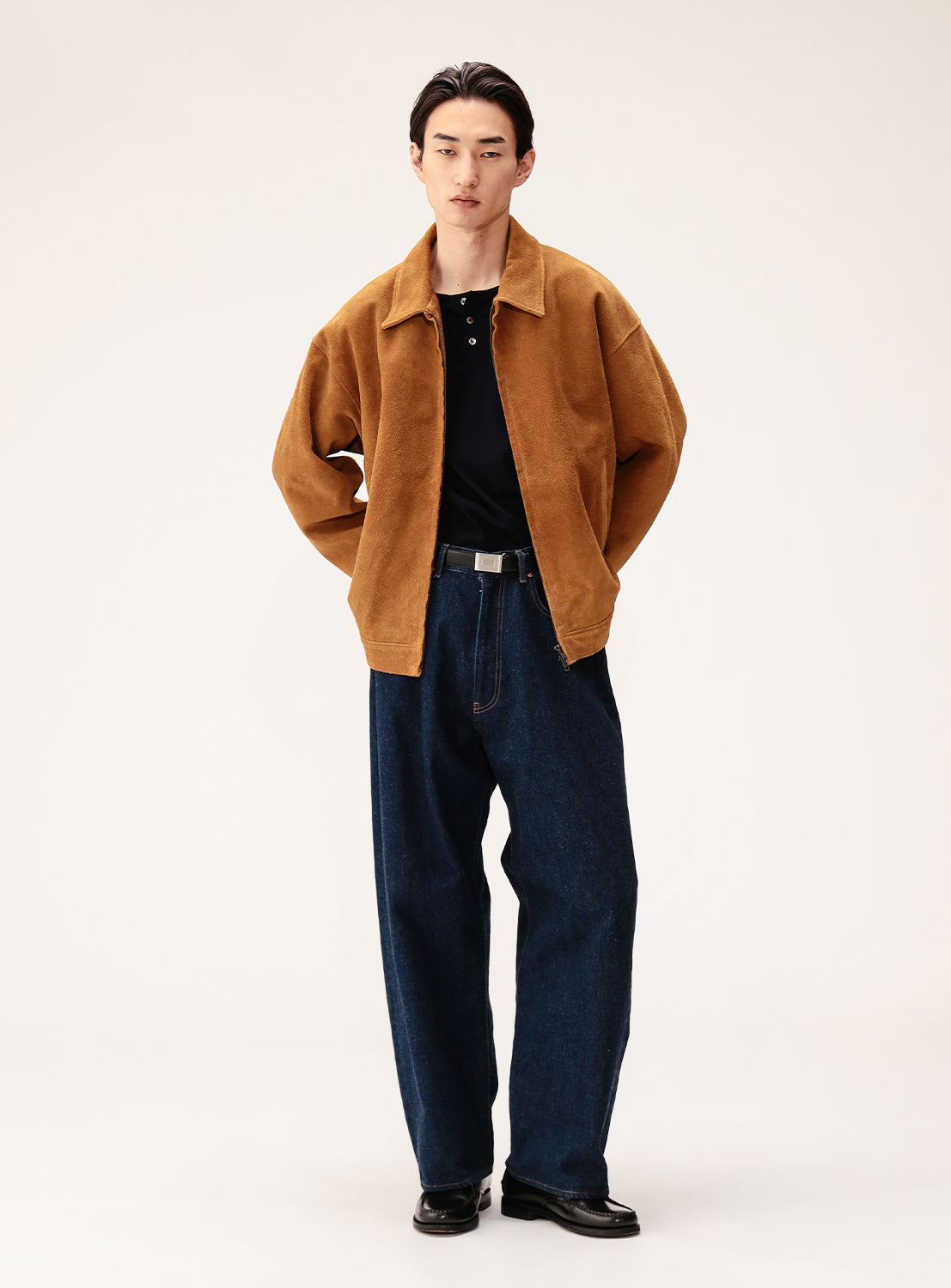 A person stands against a plain background in a vintage look, wearing BEAMS' Suede Zip-Up Blouson over a black polo, dark blue wide-leg jeans, and black shoes, hands in pockets and a neutral expression.