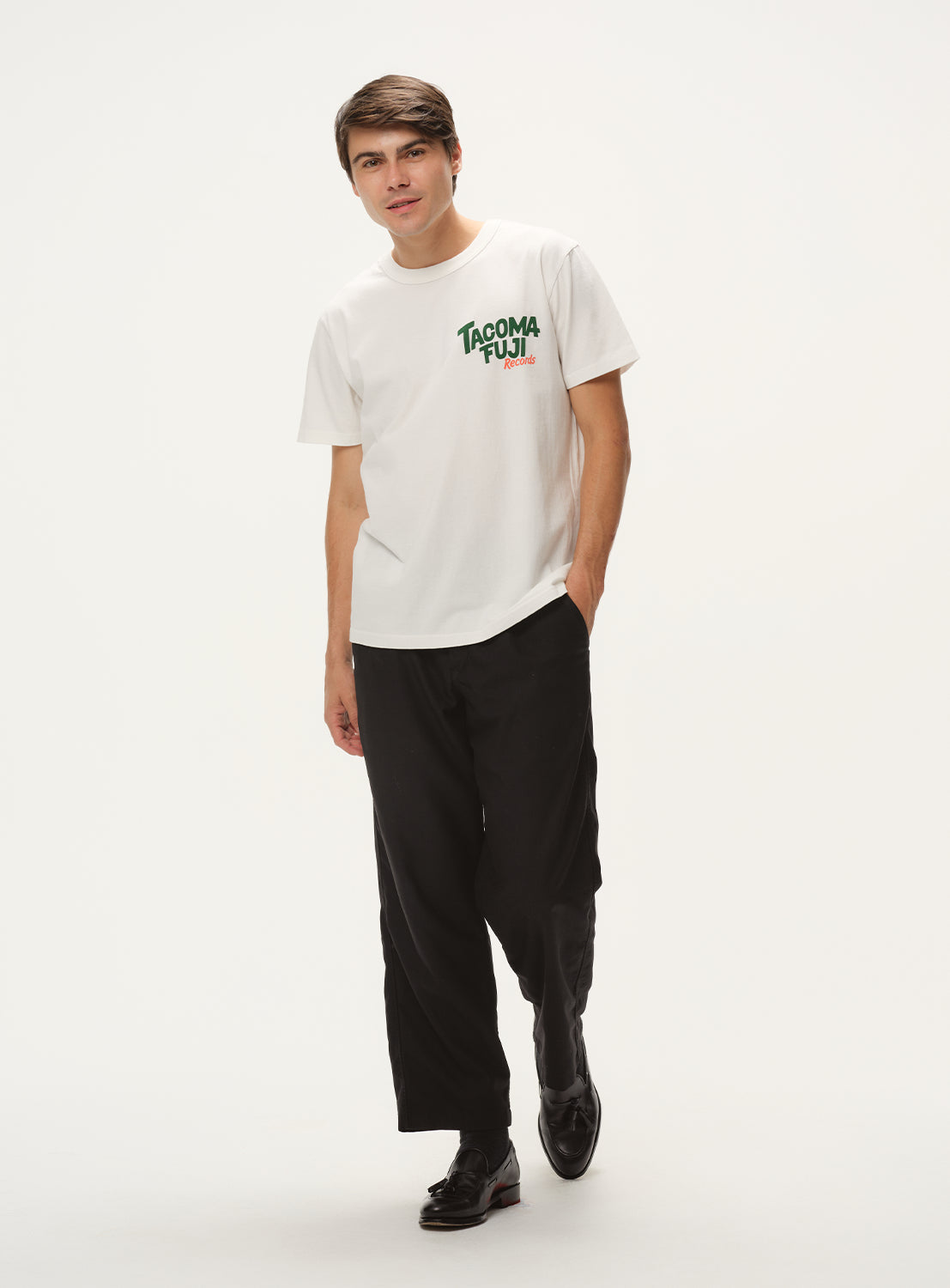 Model wearing a white t-shirt with "TACOMA FUJI Records" graphic and black shorts, full front view, displaying casual style.