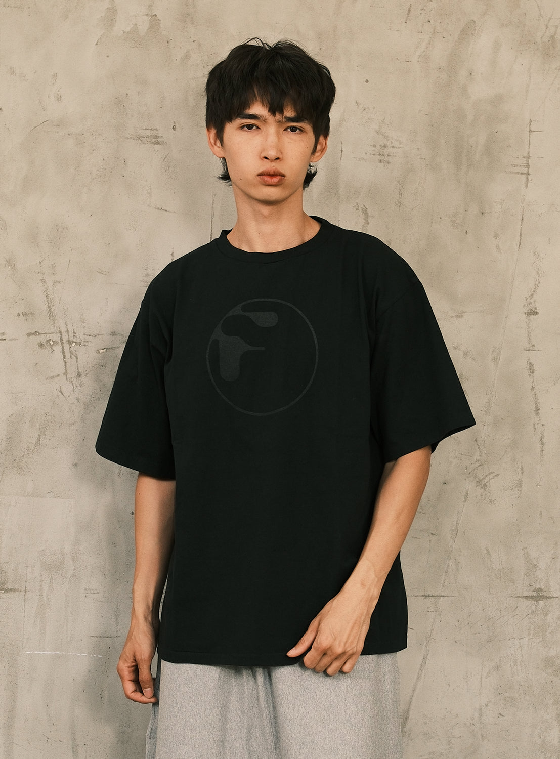 A young person with dark hair wears the F-LAGSTUF-F Symbol Logo Tee, a loose, vintage-inspired shirt crafted from heavyweight cotton jersey and featuring a subtle circular logo design. They stand against a textured gray wall, looking directly at the camera with a neutral expression.