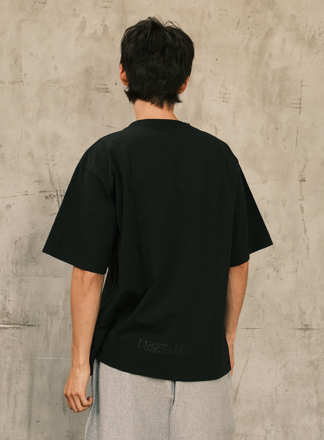A person with short dark hair stands with their back to the camera, wearing the F-LAGSTUF-F Symbol Logo Tee—an oversized black T-shirt crafted from heavyweight cotton jersey—paired with light gray pants, against a light beige textured wall.