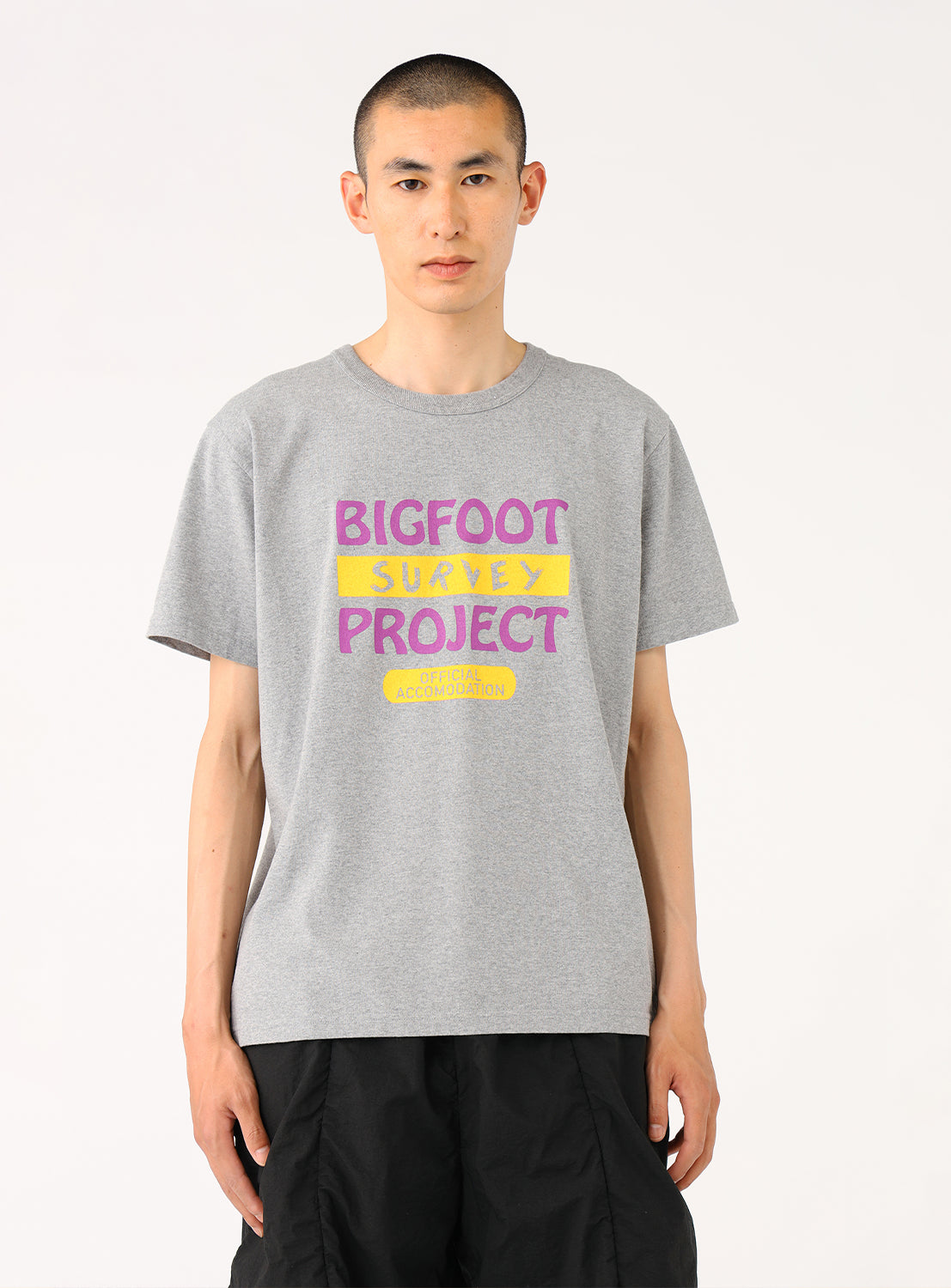 Model wearing a light grey "BIGFOOT PROJECT" t-shirt and black shorts, front view, displaying casual fit.