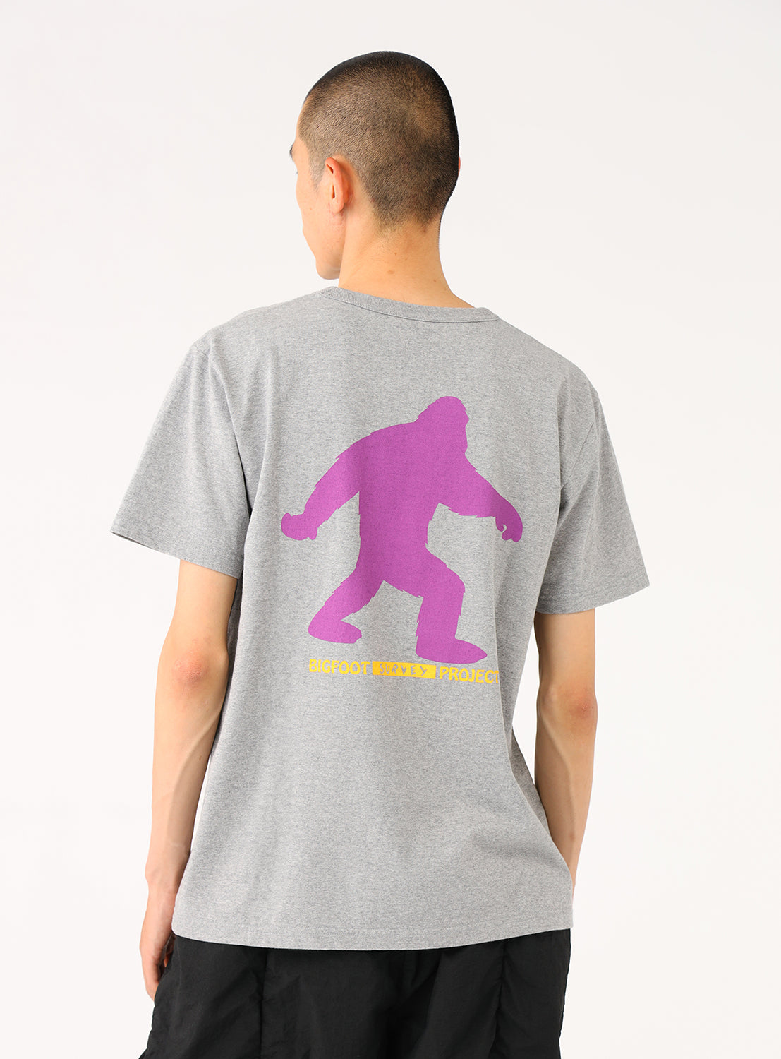 Model wearing a light grey t-shirt, back view, highlighting the purple Bigfoot silhouette graphic.