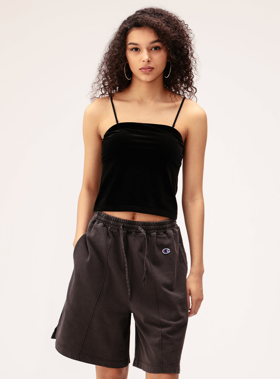 A person with curly hair models the Ray BEAMS 2-Way Convertible Velour Camisole in black, featuring built-in cups, paired with gray drawstring shorts and standing with hands in pockets against a plain light background.