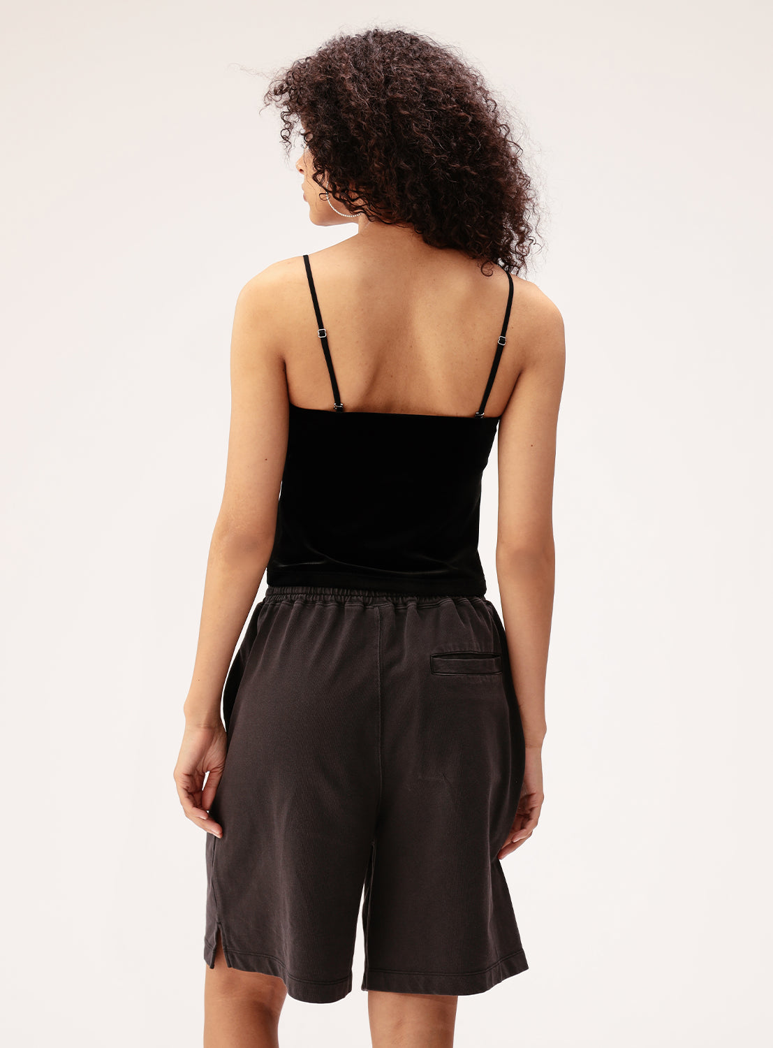 A person with curly hair is seen from behind, wearing the Ray BEAMS 2-Way Convertible Velour Camisole with removable straps and loose dark shorts, standing against a plain light background.