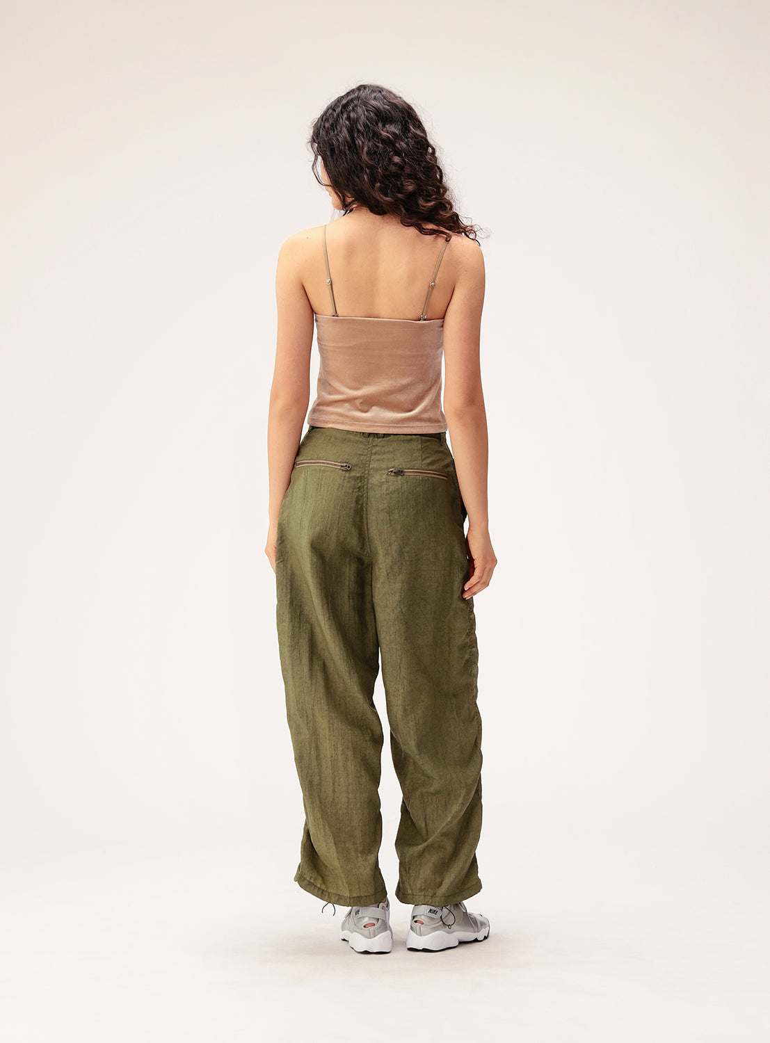 A person with long, curly hair stands facing away, wearing the Ray BEAMS 2-Way Convertible Velour Camisole in beige with removable straps, paired with loose olive green pants and white sneakers against a plain light background.