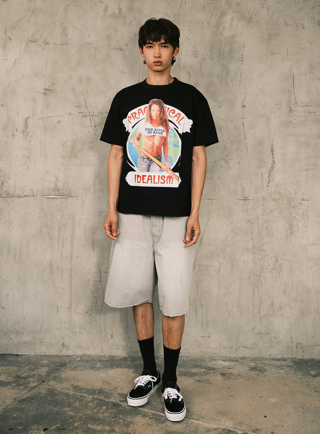 A person stands against a textured gray wall wearing BIAS DOGS’ The Rip Off Warrior Tee—a black graphic T-shirt featuring a bold photo print—paired with light gray knee-length shorts, black socks, and black sneakers for a 1990s street culture-inspired look.