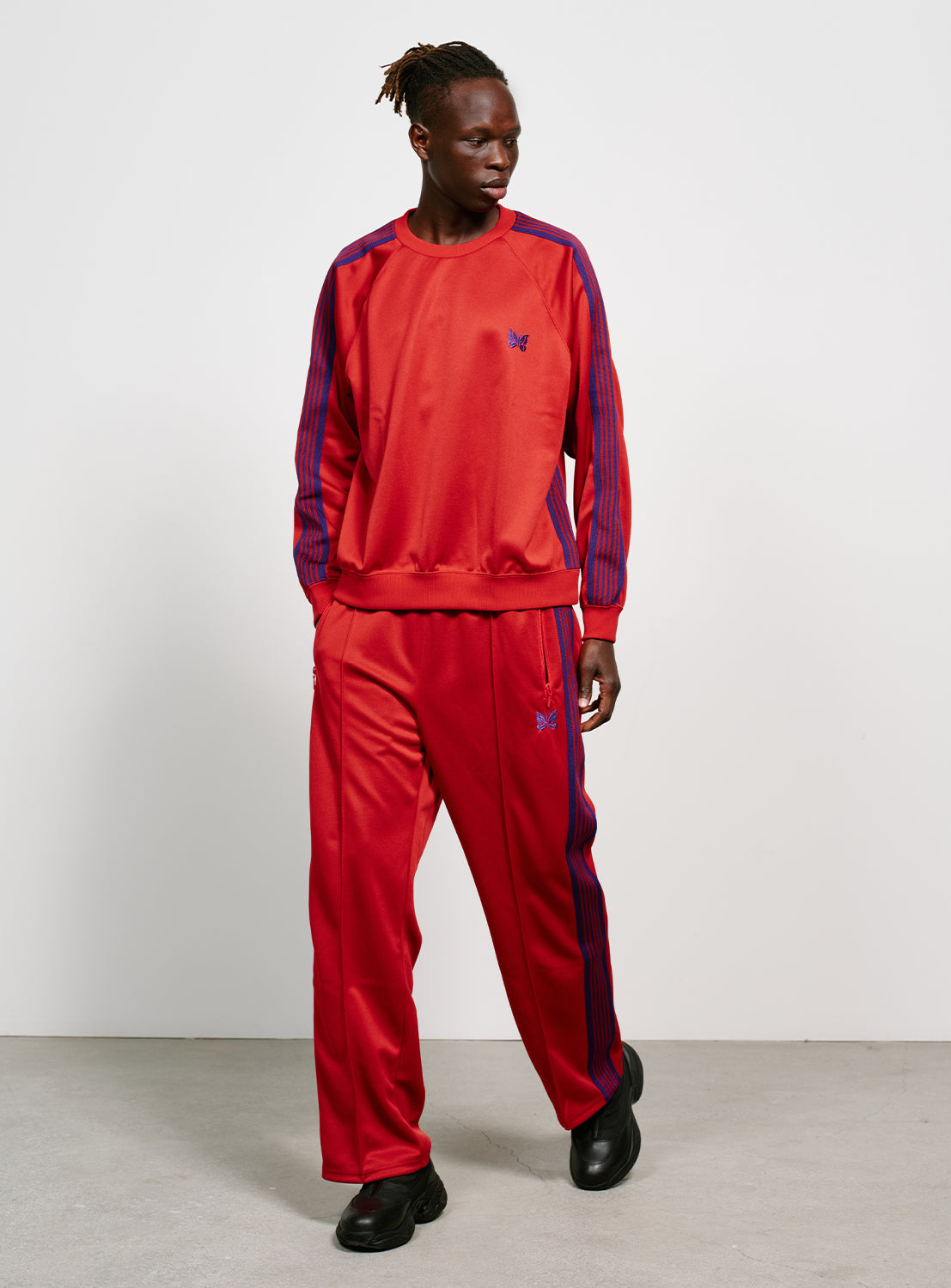 Model wearing a red track pants with blue/purple stripes on the side, two zipper pockets and a purple embroidered butterfly. 