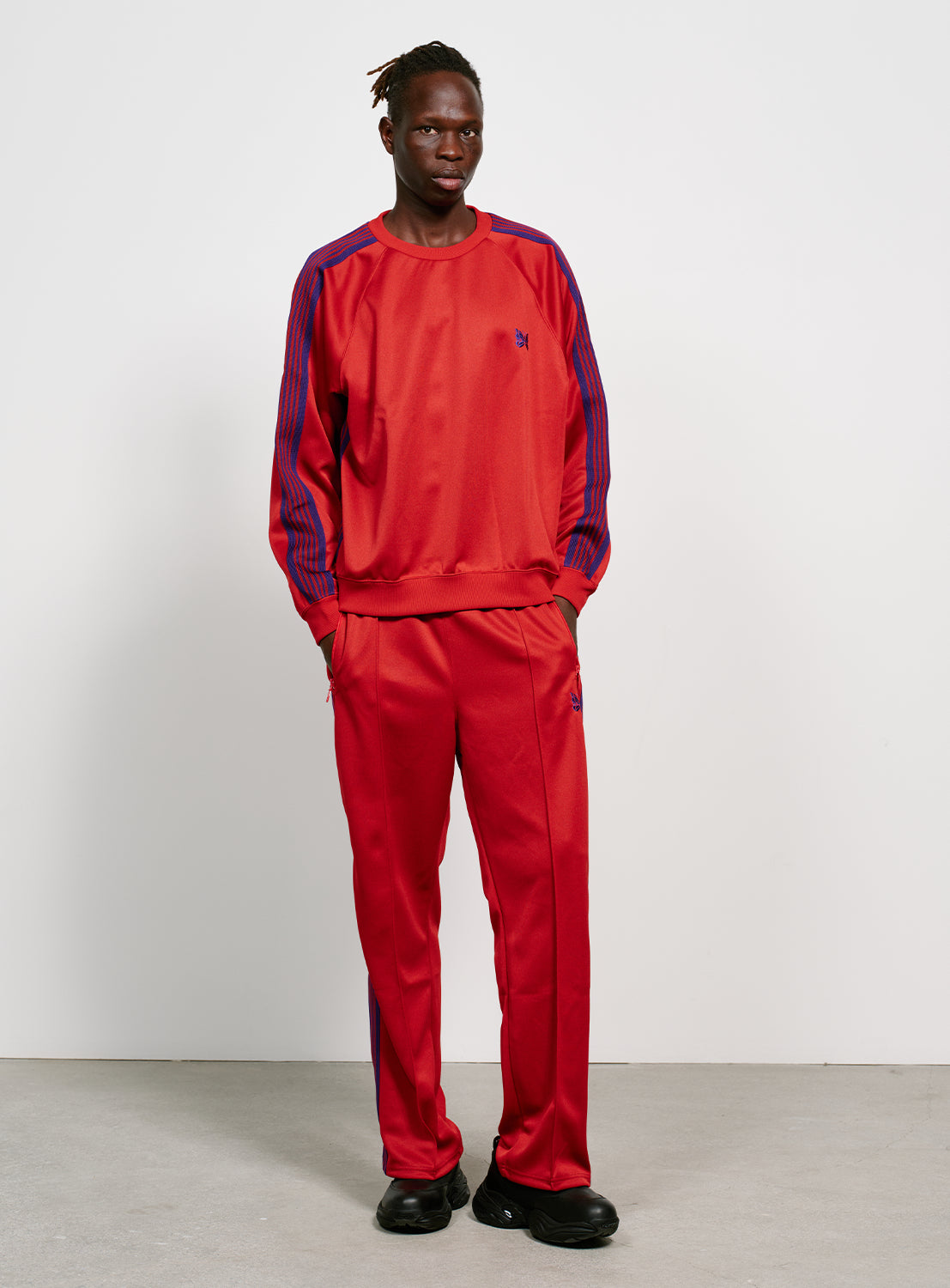 Model wearing red track pants with blue/purple stripes on the side, two zipper pockets and a purple embroidered butterfly. 