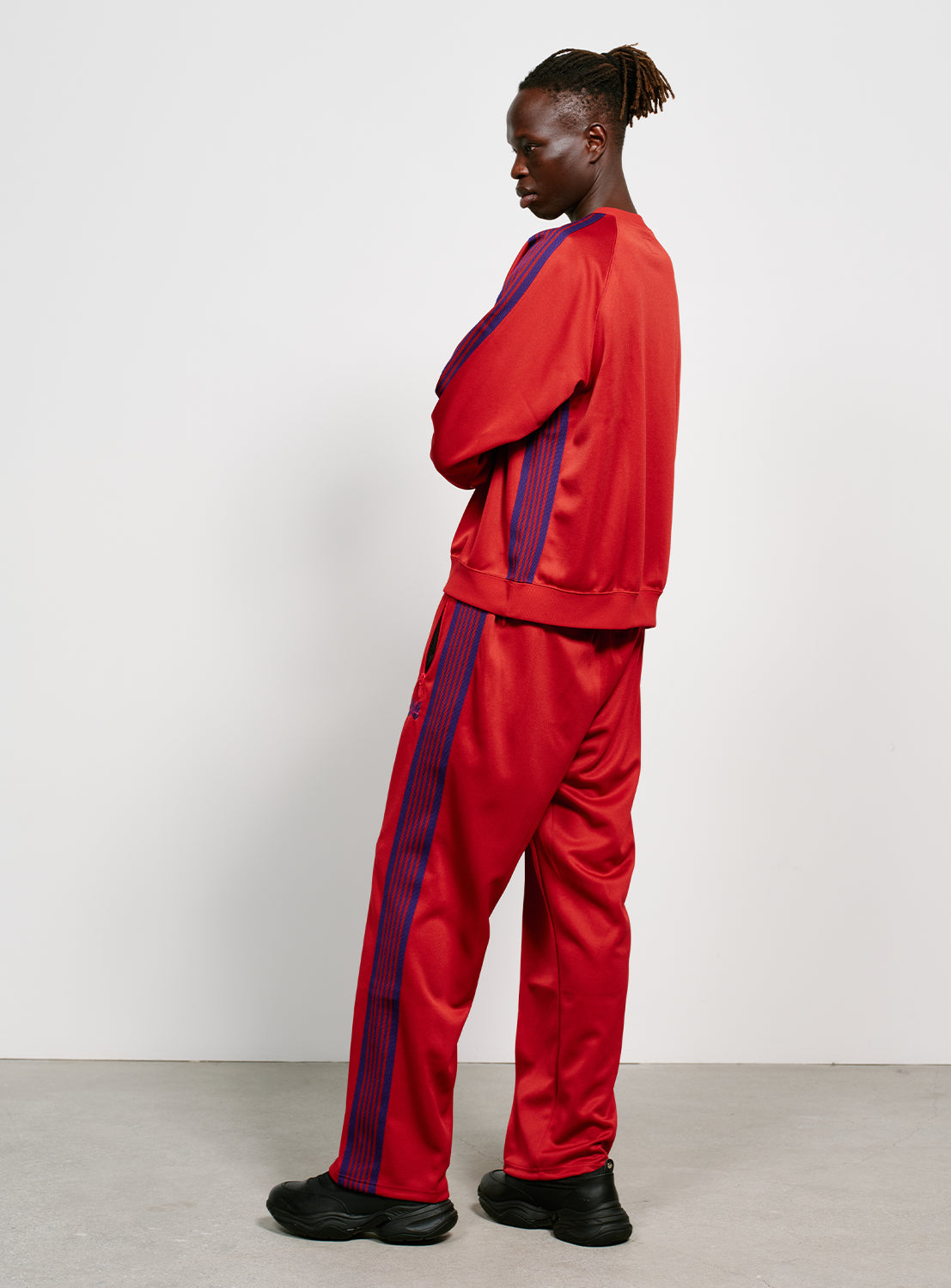 Side view of a model wearing a red track pants with blue/purple stripes on the side, two zipper pockets and a purple embroidered butterfly. 