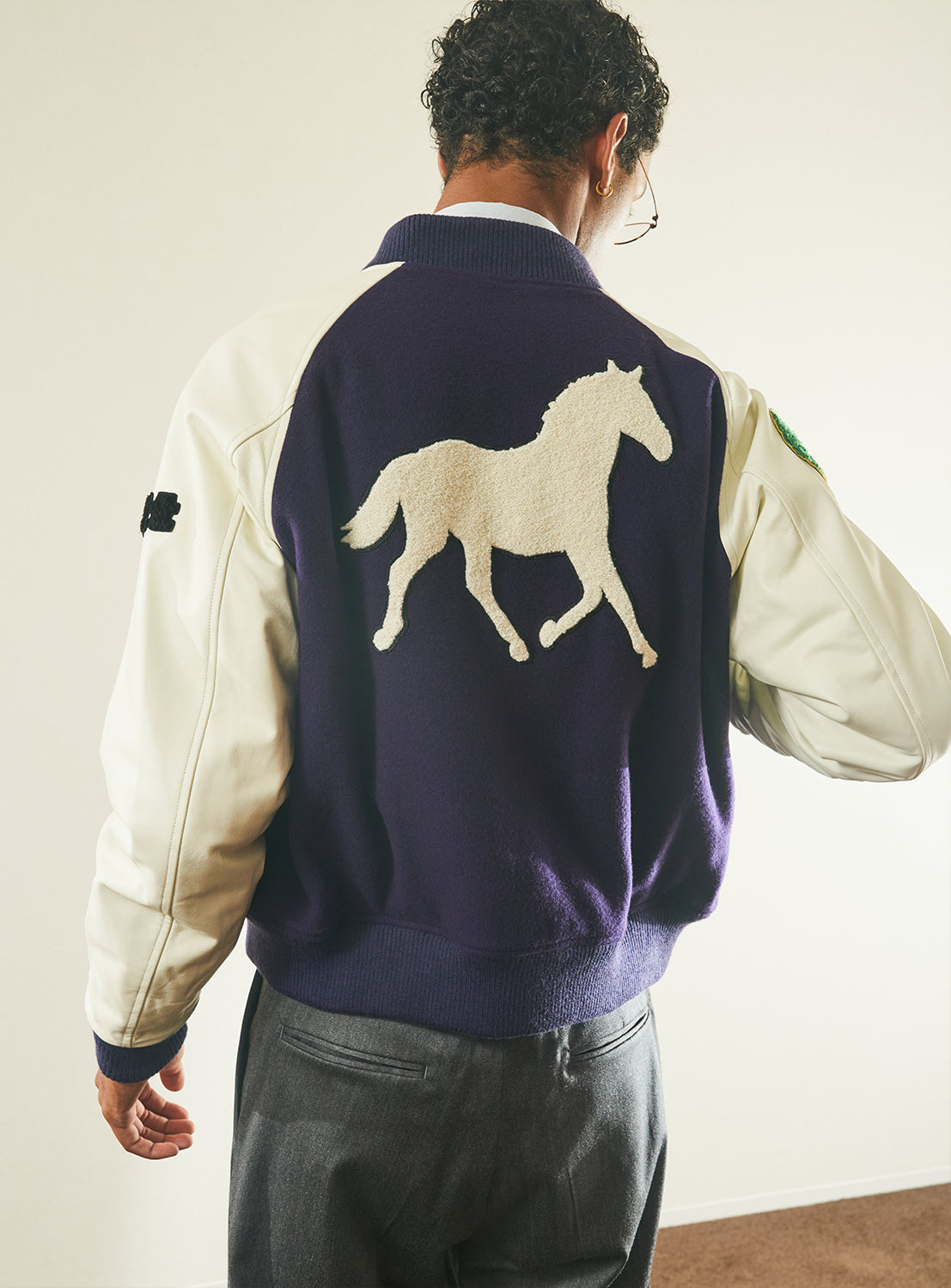 A person is standing indoors, facing away from the camera, wearing the TTT MSW Varsity Patch Jacket—a modern fit piece in purple and white with a distinctive white horse design on the back, crafted from soft lamb wool.