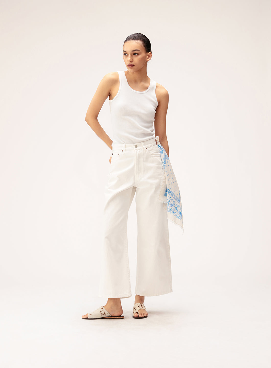 A woman stands against a plain background wearing a white tank top, wide-leg white pants styled with a blue-patterned scarf at the waist, and white sandals. She accessorizes her look with the BEAMS BOY 2-Way Floral Bandana worn as a hair accessory, gazing to the side with her hands in her pockets.