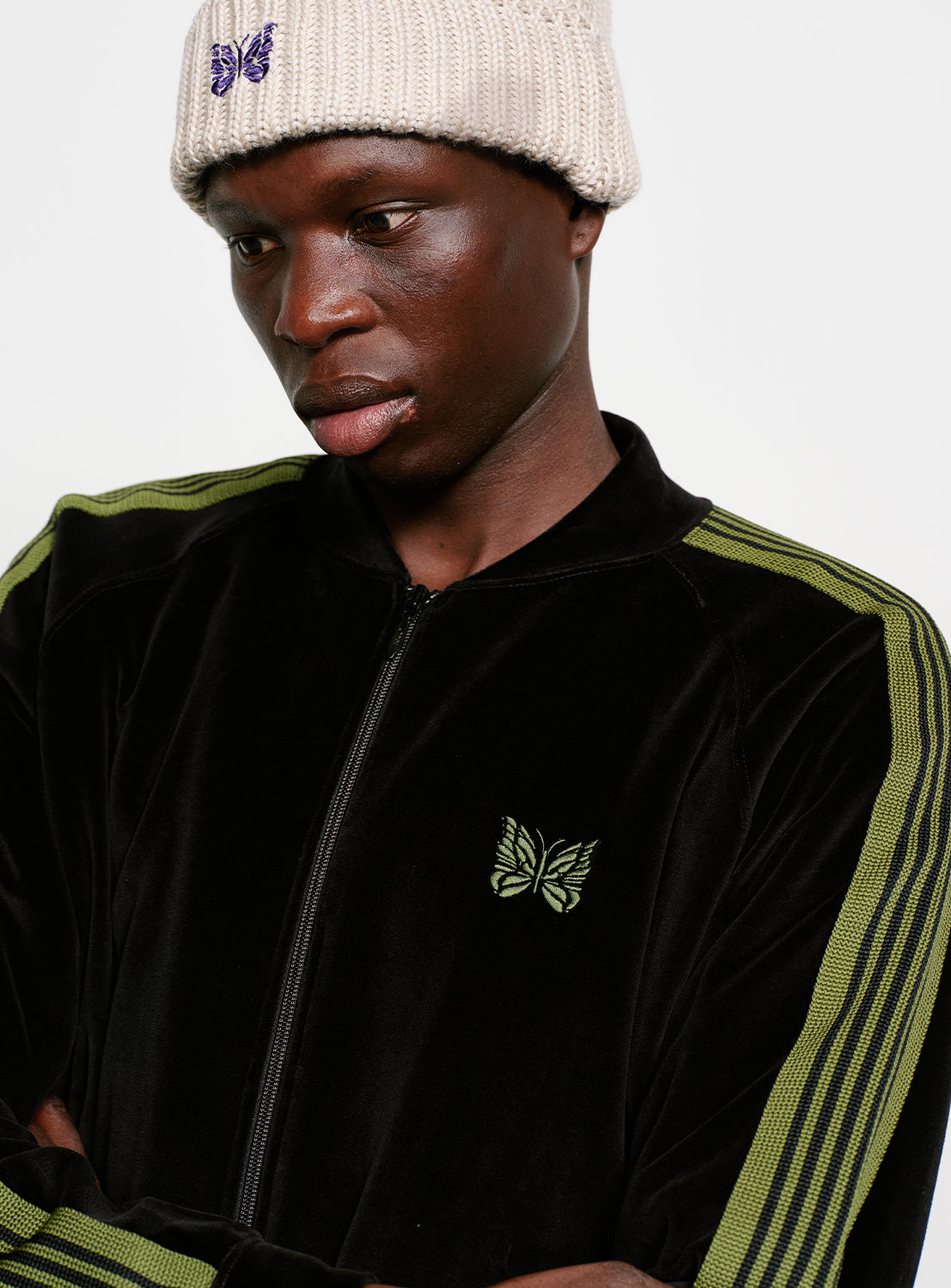 Model wearing a black tracksuit with green stripes and a beige beanie, with butterfly details. 