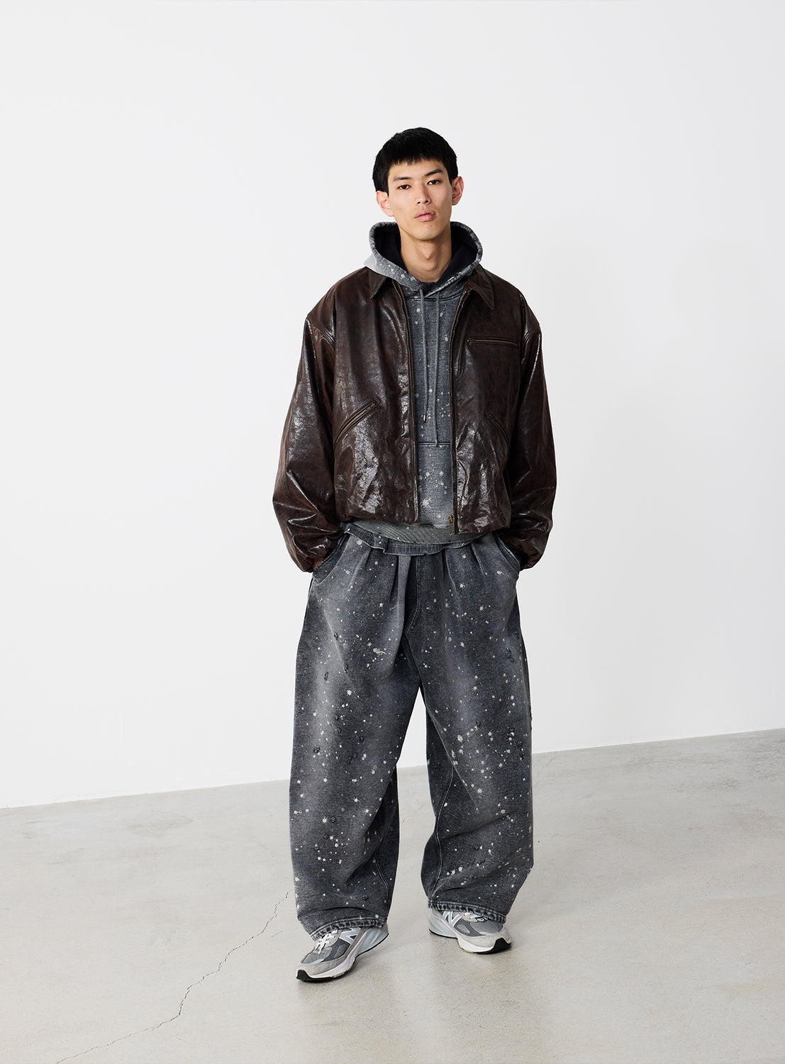 A young man stands indoors against a plain white wall, wearing a dark brown leather jacket over the BEAMS Wide Short Fit Distressed Heavyweight Hoodie, paired with wide-legged gray splatter-patterned pants and gray sneakers.