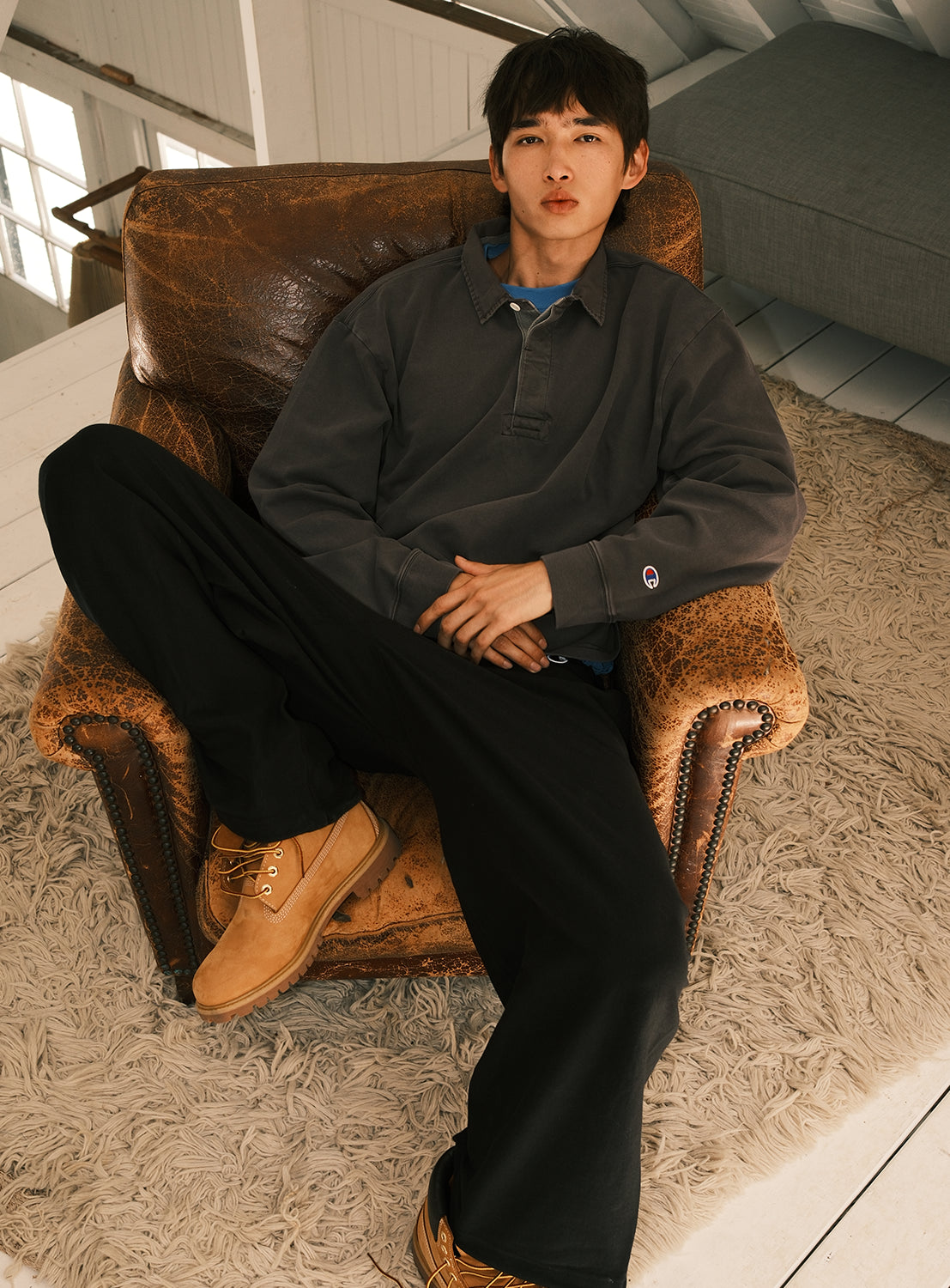 A person with straight dark hair sits relaxed in a worn brown armchair, wearing the [Exclusive] BEAMS x Champion Heavyweight Pigment-Dyed Rugby Shirt by CHAMPION, paired with black pants and tan boots, hands resting on their stomach atop a textured beige rug in a cozy, sunlit room.
