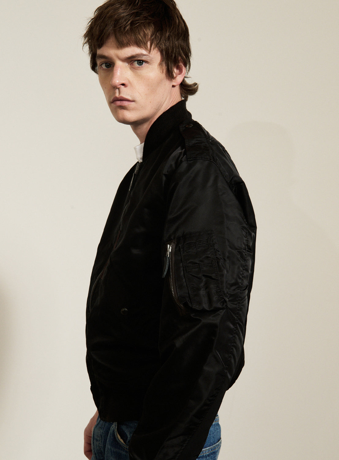 A young man with short brown hair, wearing a black BUZZ RICKSON’s William Gibson Collection L-2B jacket made in Japan and blue jeans, stands sideways, looking over his shoulder at the camera against a plain, light-colored background.