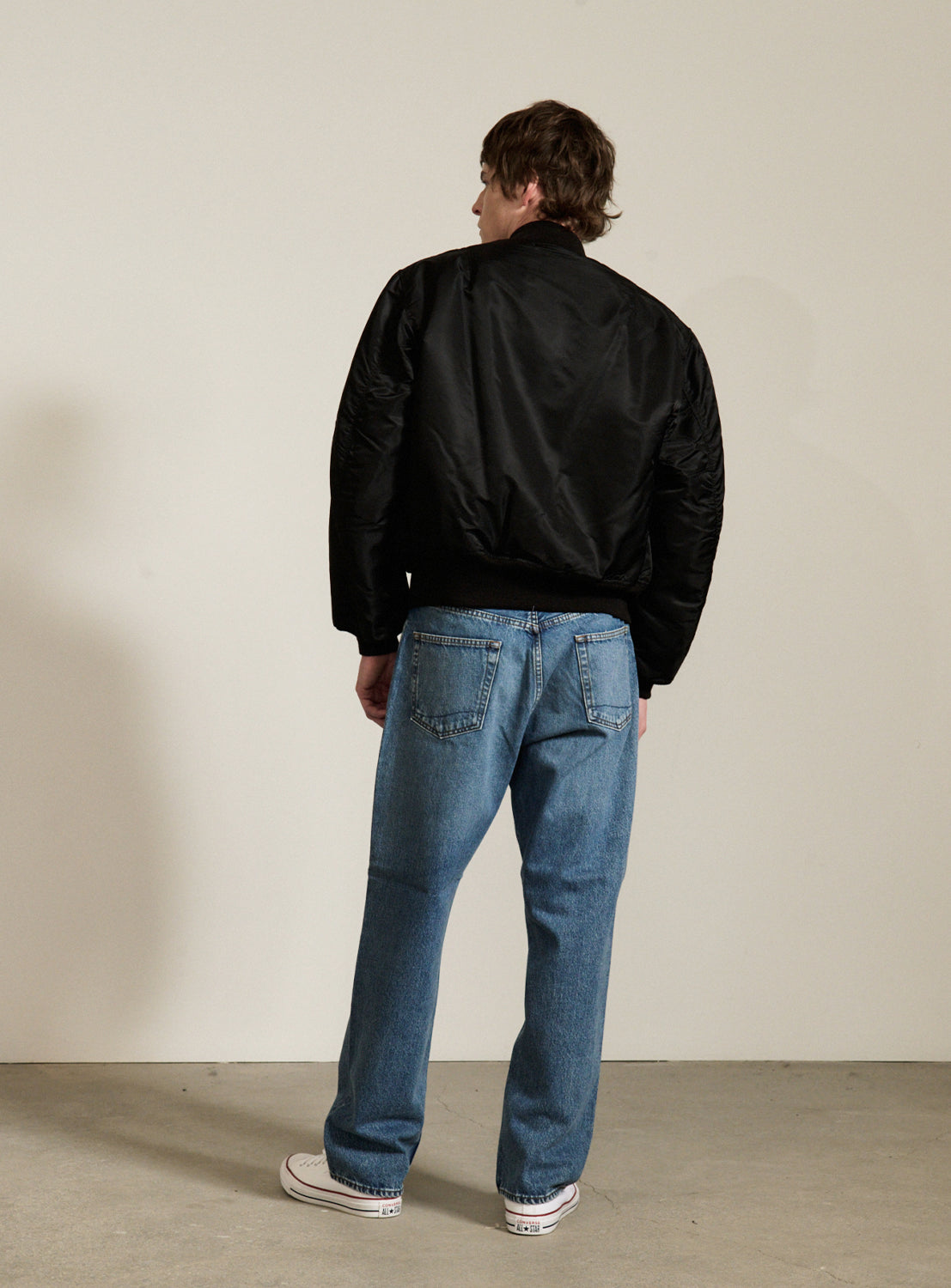 A person stands indoors with their back to the camera, wearing a black BUZZ RICKSON’s William Gibson Collection MA-1 jacket, blue jeans, and white sneakers. The background features a plain white wall and a gray floor.