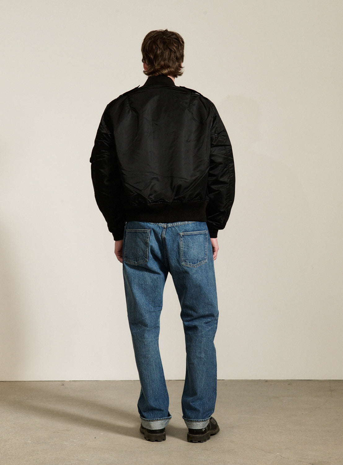 A person stands facing a blank wall, wearing a black BUZZ RICKSON’s William Gibson Collection L-2B jacket, blue jeans, and black shoes. The floor is concrete, and the person's hands are at their sides. This classic bomber jacket is proudly Made in Japan.