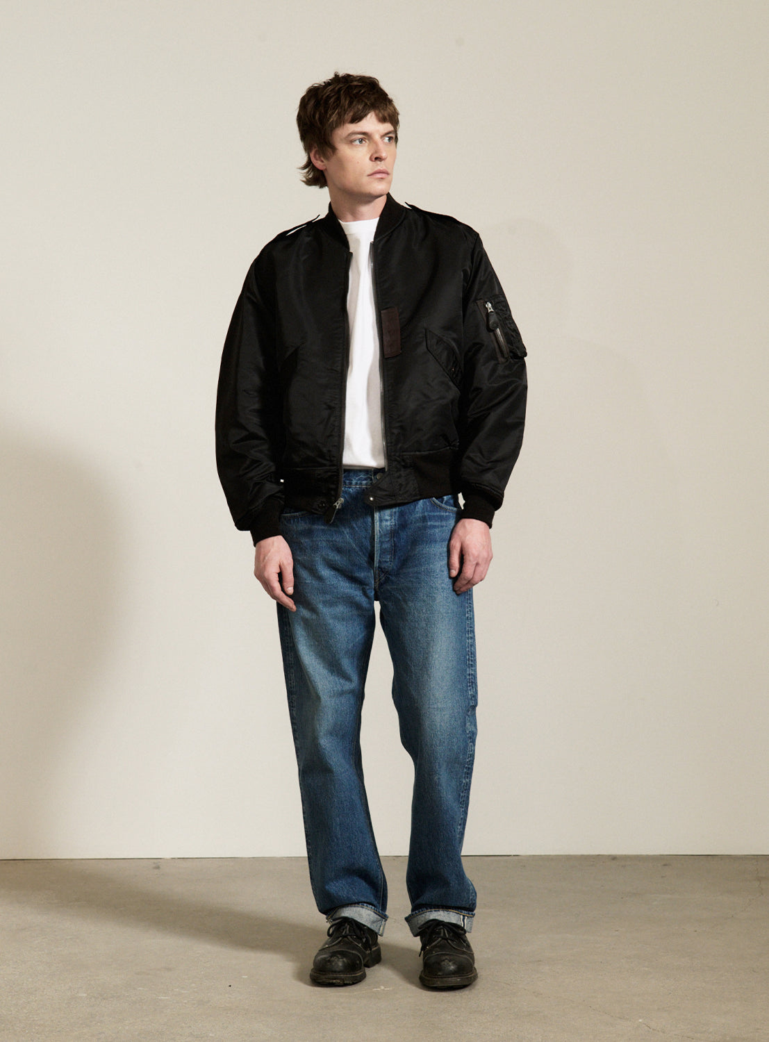 A person stands indoors wearing a BUZZ RICKSON’s William Gibson Collection L-2B jacket, paired with a white t-shirt, blue jeans, and black shoes, looking slightly to the right against a plain light-colored wall. This classic bomber jacket from BUZZ RICKSON’s is crafted in Japan and showcases exceptional quality and timeless style.