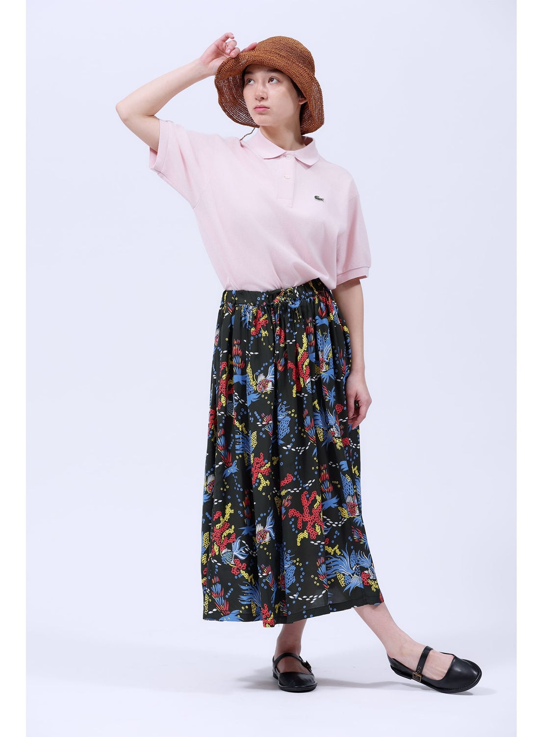 Wearing a brown sun hat, light pink polo, and the [Exclusive] SUN SURF x BEAMS BOY Aloha Gathered Long Skirt by SUN SURF, a person stands against a white background, one hand on their hat and the other resting by their side.