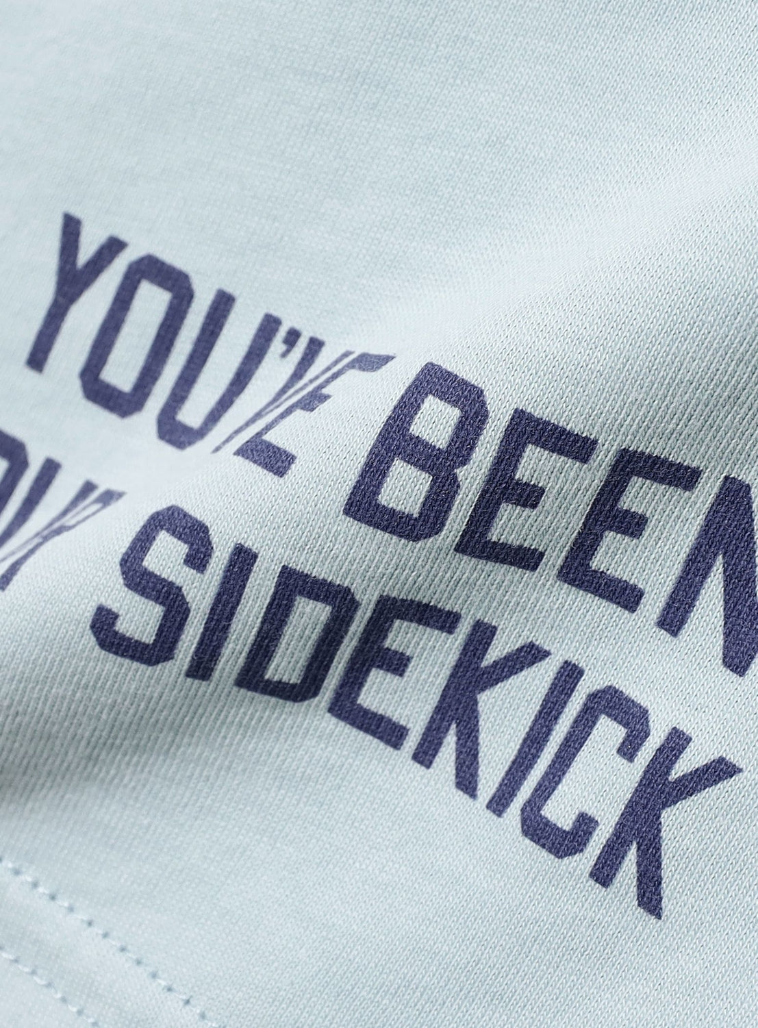 Close-up of light blue fabric from the BEAMS BOY Big Logo Long-Sleeve T-Shirt, featuring partially visible navy text that reads “YOUVE BEEN” and “SIDEKICK.” The slightly wrinkled material subtly distorts portions of the text. This relaxed fit tee by BEAMS BOY highlights both comfort and style in its thoughtful design.