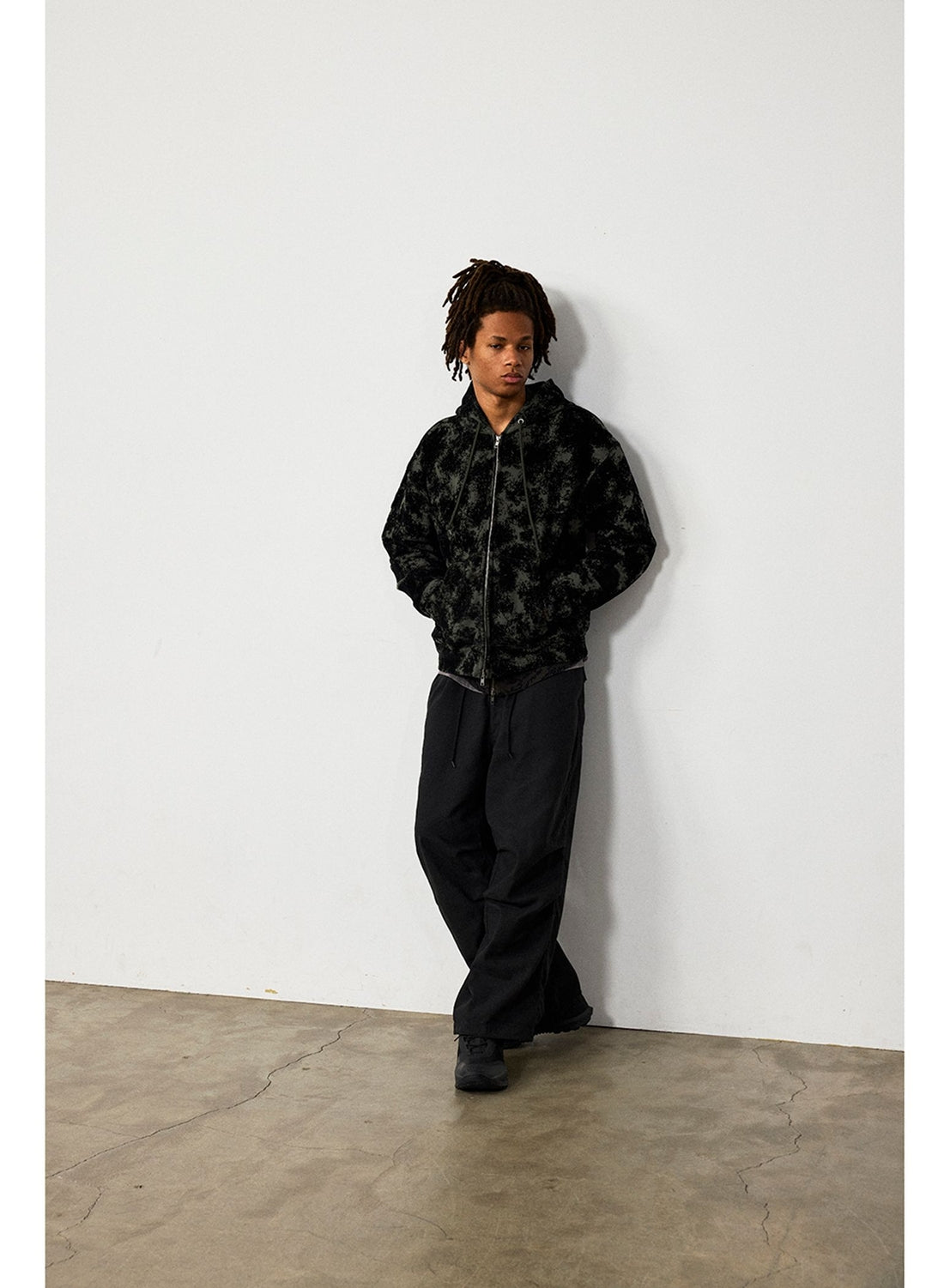 A person wearing a BEAMS Flocked Planet Motif Full-Zip Hoodie in black with a faint textured print and loose-fitting black pants, leaning against a white wall.